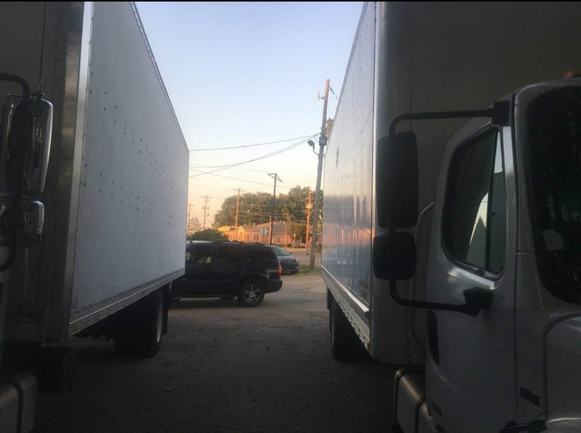 Two white delivery trucks parked facing each other with their cabs on the right side of the image, and a black SUV parked in the background near a street with power lines and trees at sunset.