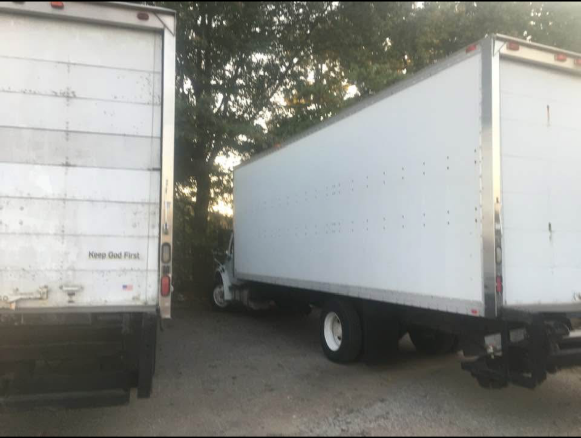 Two white box trucks parked next to each other on a dirt surface, with trees in the background. The left truck has a sticker that says 'Keep God First'.