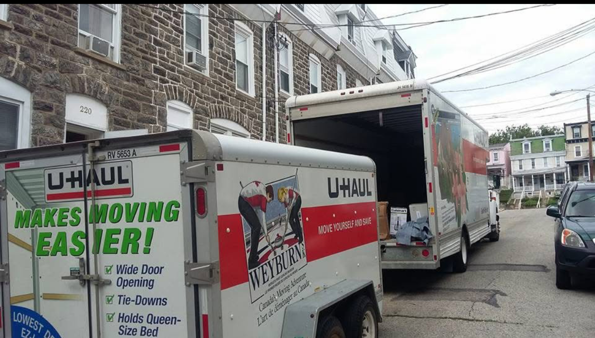 Two U-Haul trucks parked on a residential street, with the smaller one in front. The smaller truck has advertisements promoting moving services, including wide door opening, tie-downs, and Queen-size bed holds. The larger truck is open, revealing box