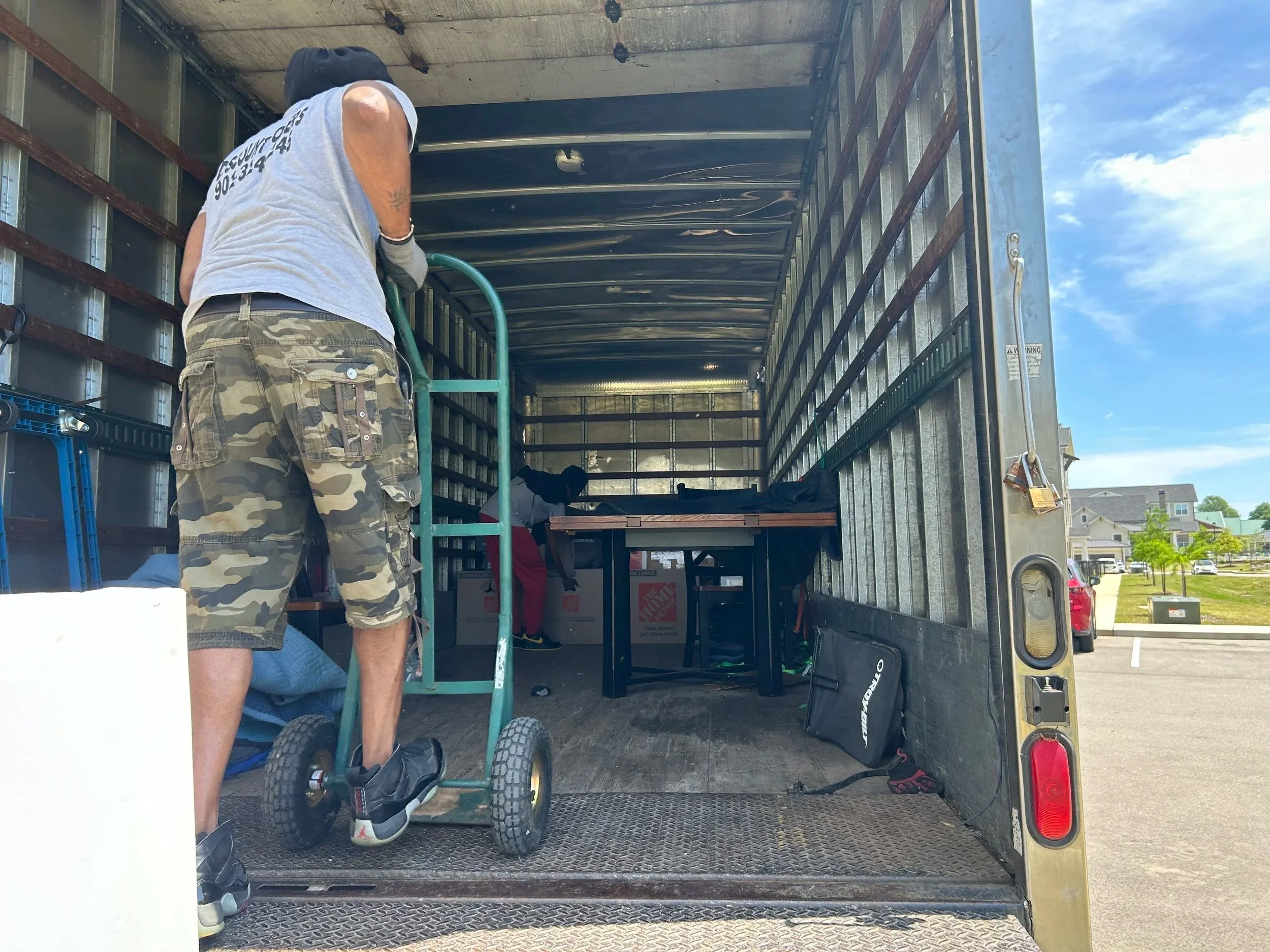 A person wearing camouflage shorts and a gray t-shirt, standing on a hand truck inside a truck, with another person working in the back of the truck, and a parking lot with houses and cars visible outside.