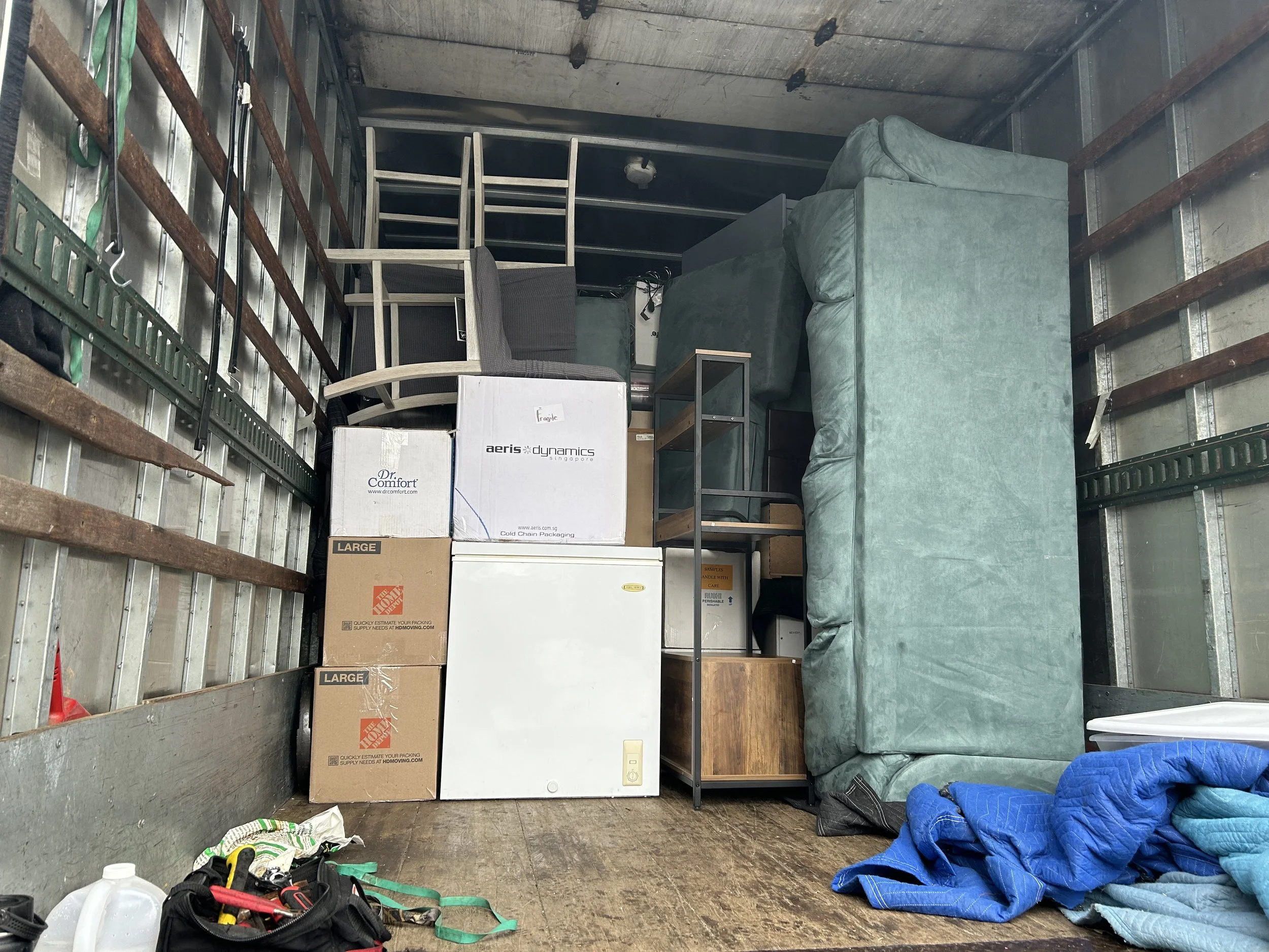 Interior of a moving truck filled with furniture, boxes, and household items including a green sofa, wooden and metal furniture, cardboard boxes, some labeled with brand names, and various tools and objects scattered on the floor.