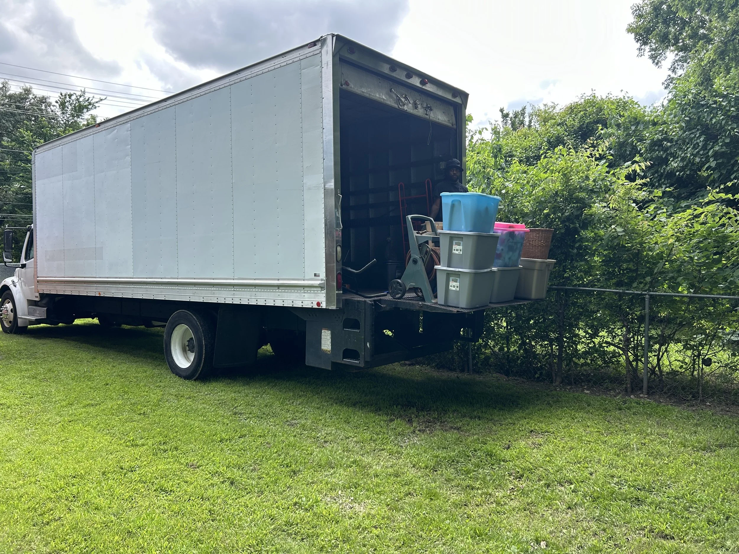 A moving truck parked on grass with an open back door filled with various plastic storage bins and household items. A man is partially visible inside the truck.