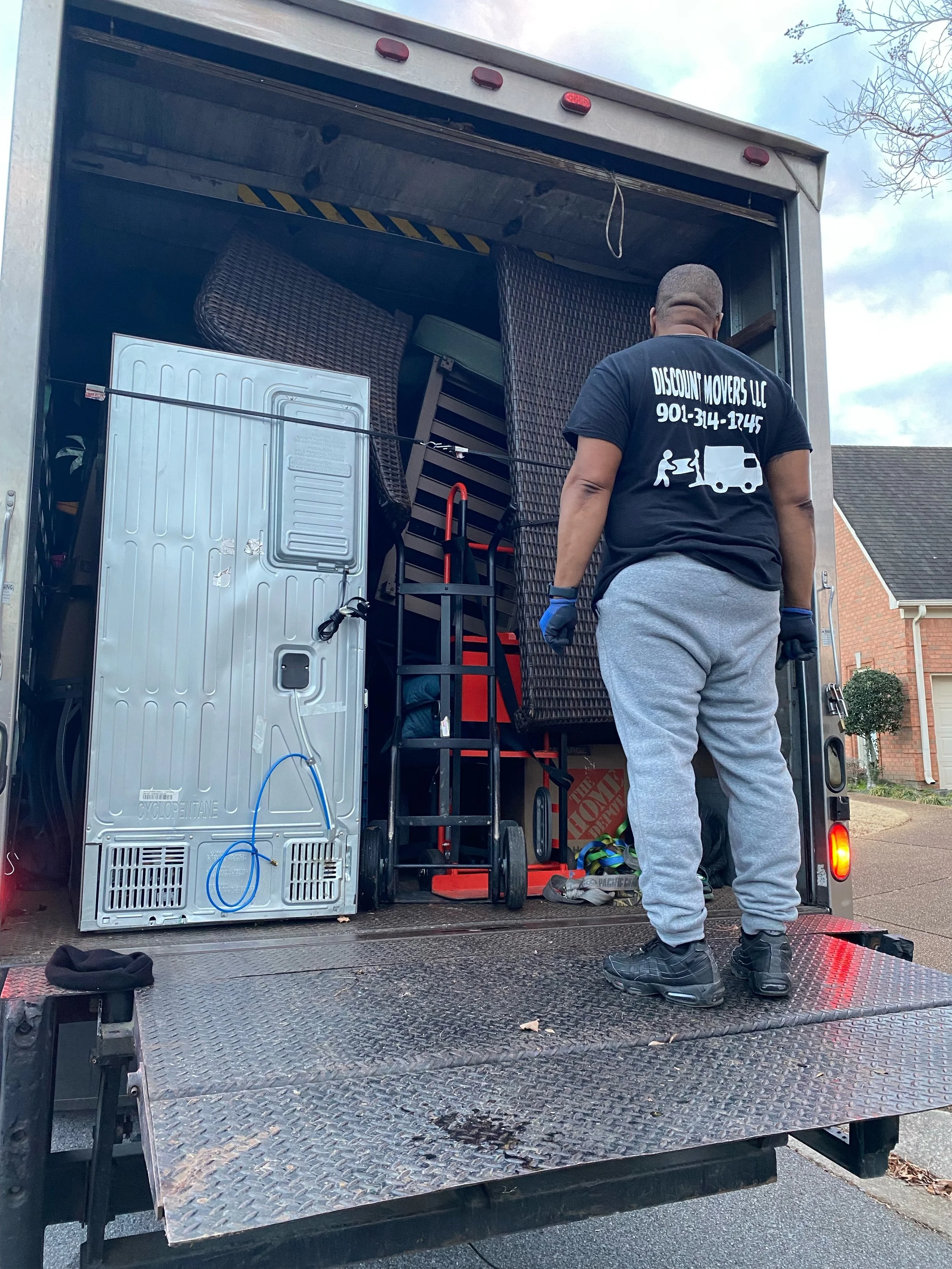 Man loading furniture and appliances into a moving truck, with furniture stacked inside and various items on the truck's platform, in a residential neighborhood with brick houses and a cloudy sky.