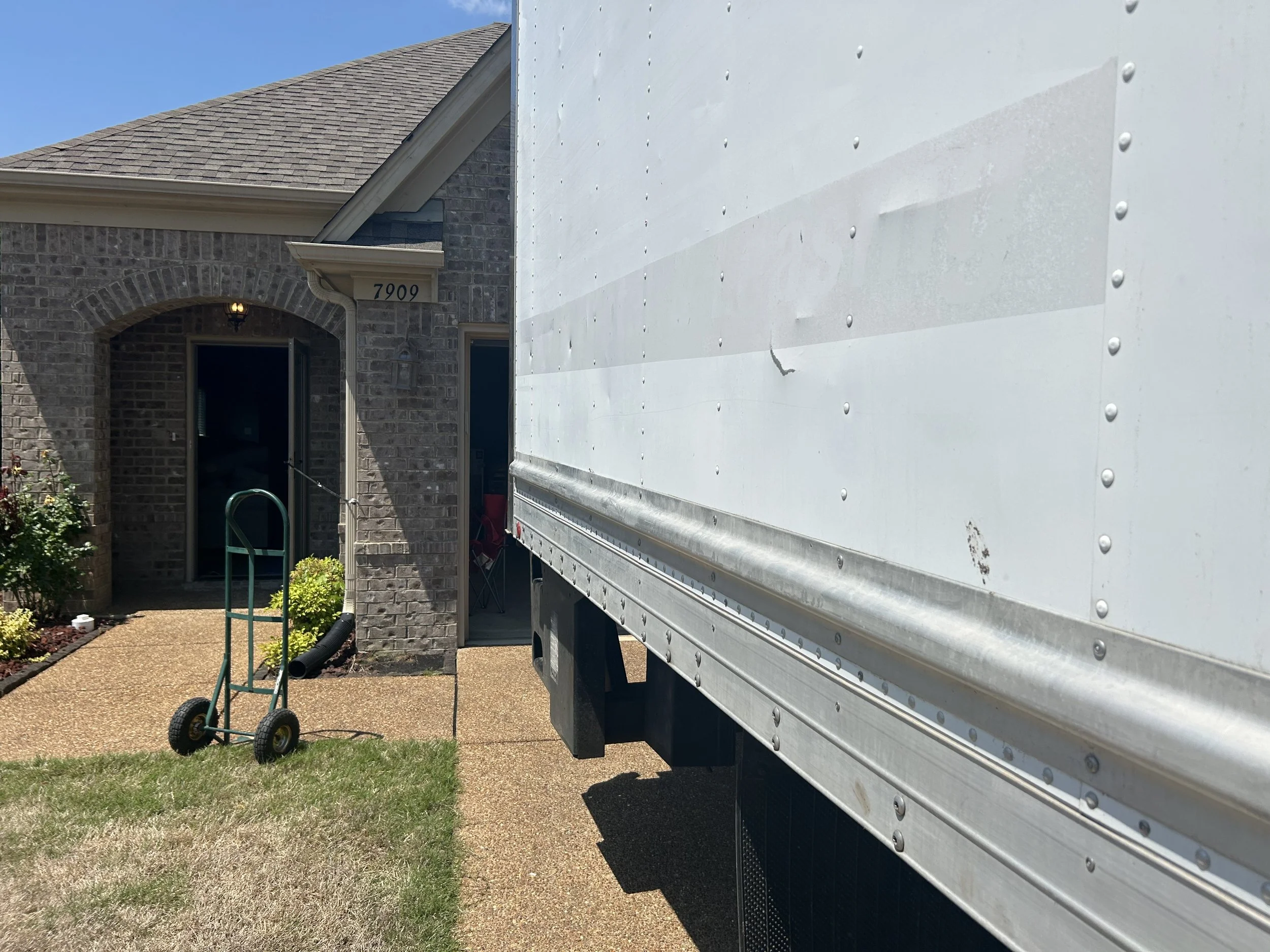 View of a house with a brick facade and shingled roof, with a partial view of a delivery truck and a hand truck in the front yard.