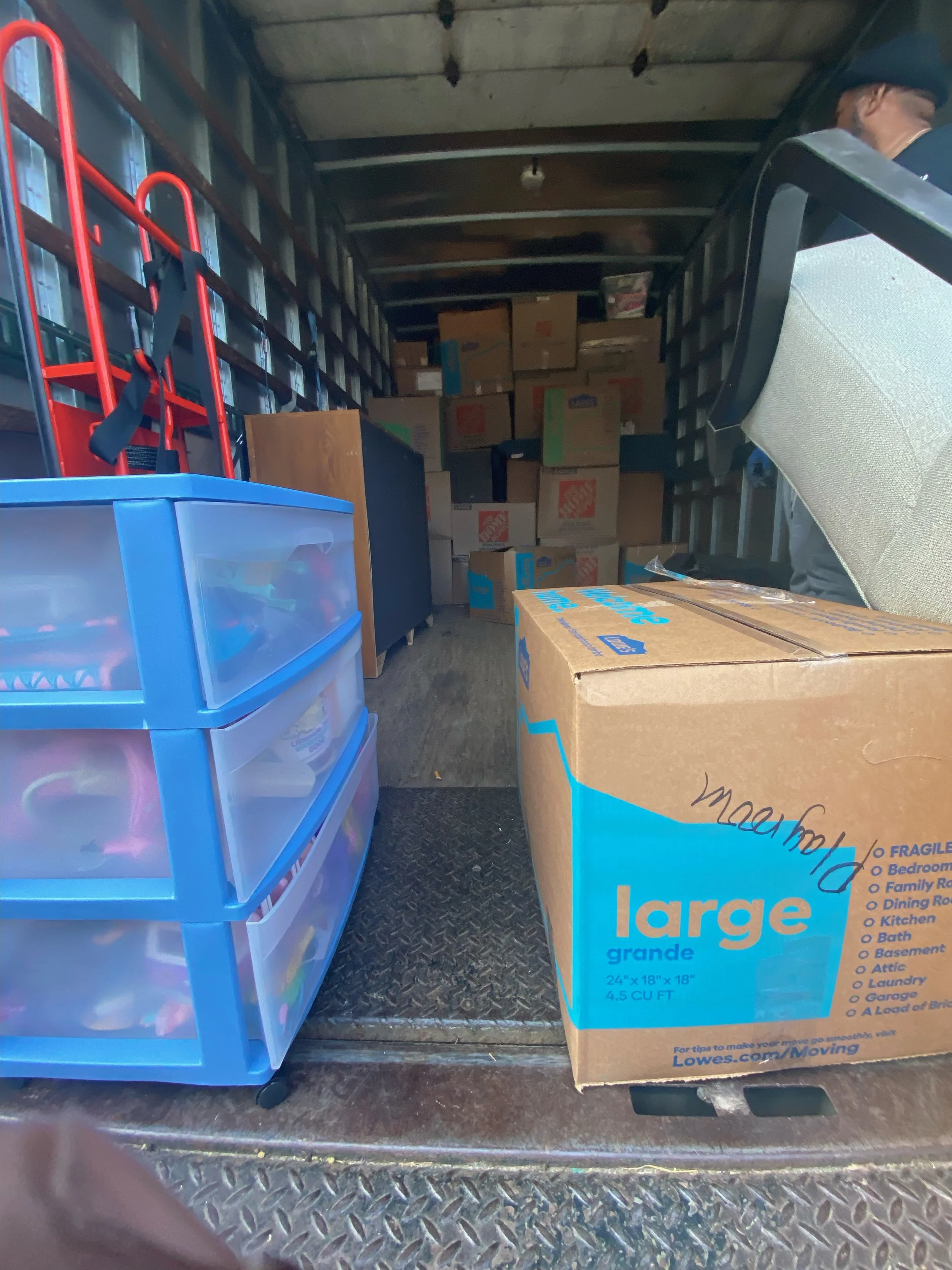 The image shows a large cardboard box labeled 'large' and measuring 24x18x18 inches, positioned on a metallic surface, with a plastic storage container filled with items and a red hand truck nearby, inside a vehicle or storage space.