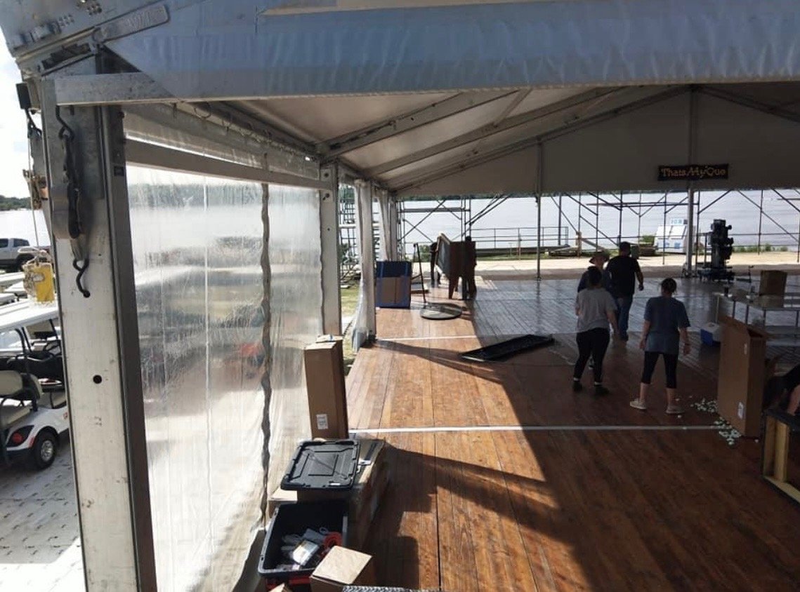 People setting up a stage under a white tent with a wooden floor, overlooking a body of water.