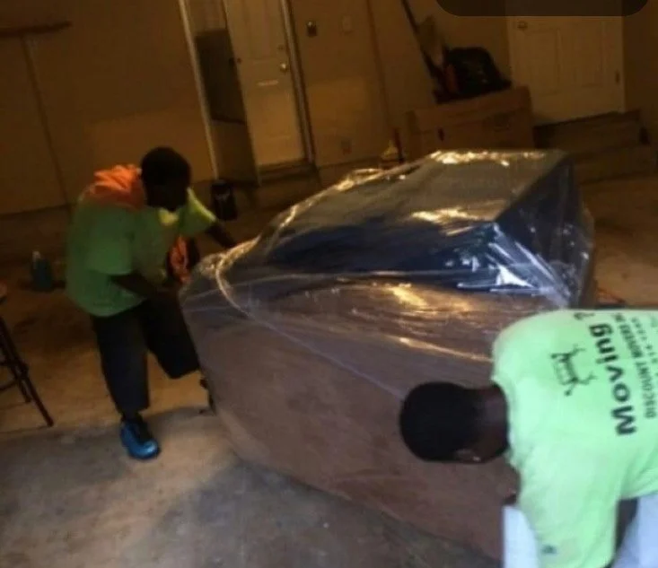 Two boys wrapping a large piece of furniture, possibly a couch, with plastic wrap in an indoor setting.