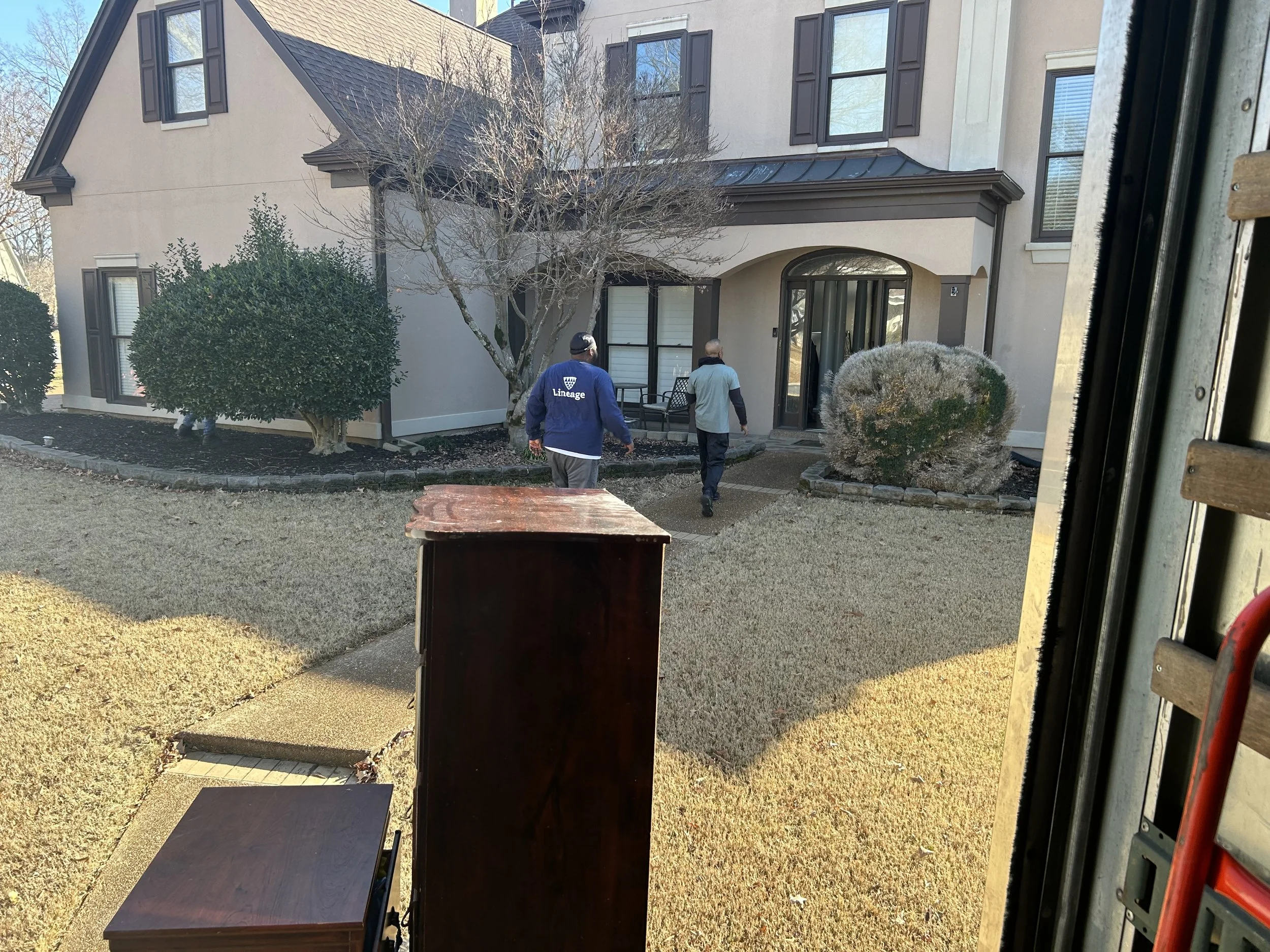 View from inside a moving vehicle showing a house with several trees and bushes in front. Two people are walking towards the house, one wearing a blue jacket with the word 'Lineage' on the back.