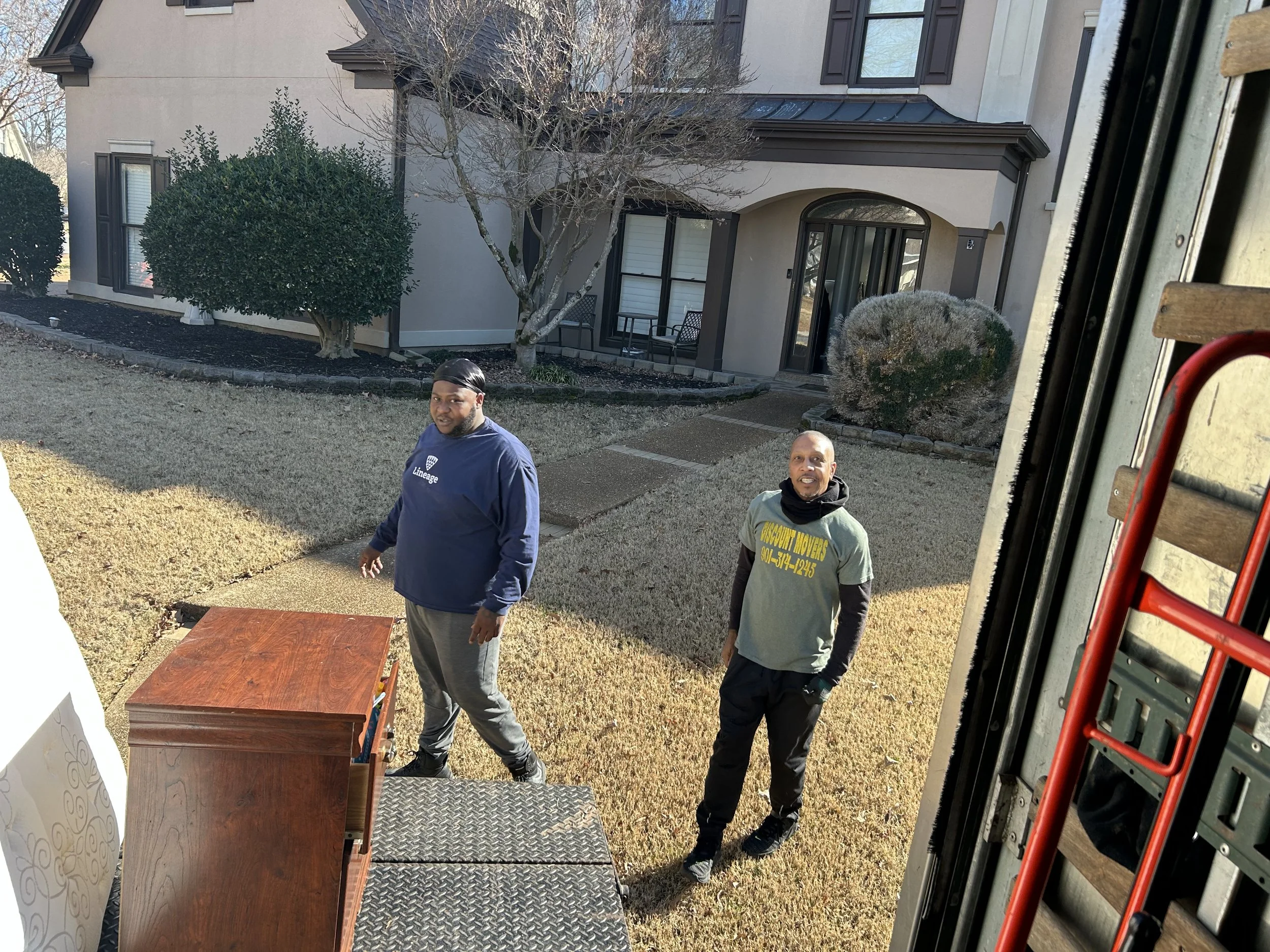 Three men standing outdoors near a house with a tree, bushes, and a porch, during daytime.
