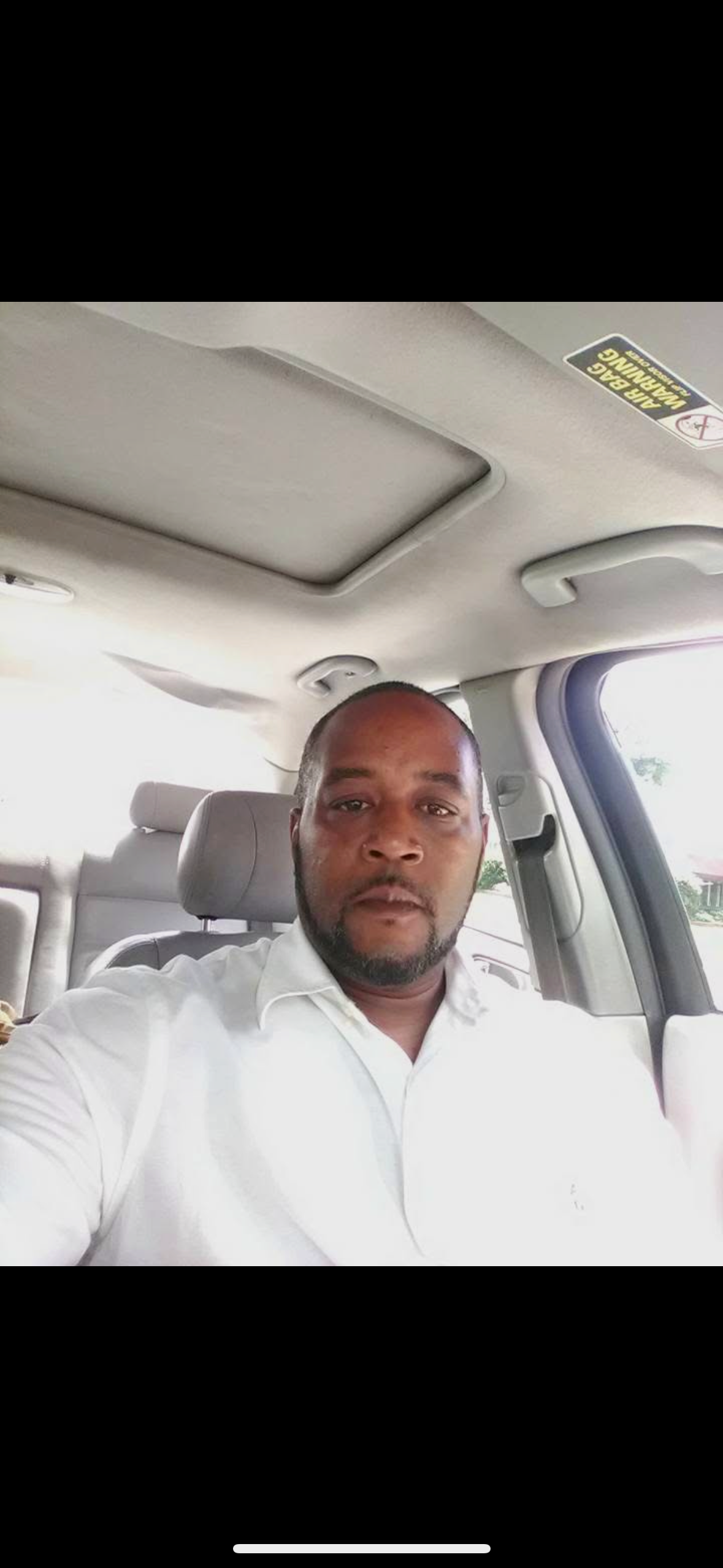 A man with a beard and mustache taking a selfie inside a vehicle, wearing a white shirt, with car interior and window visible in the background.