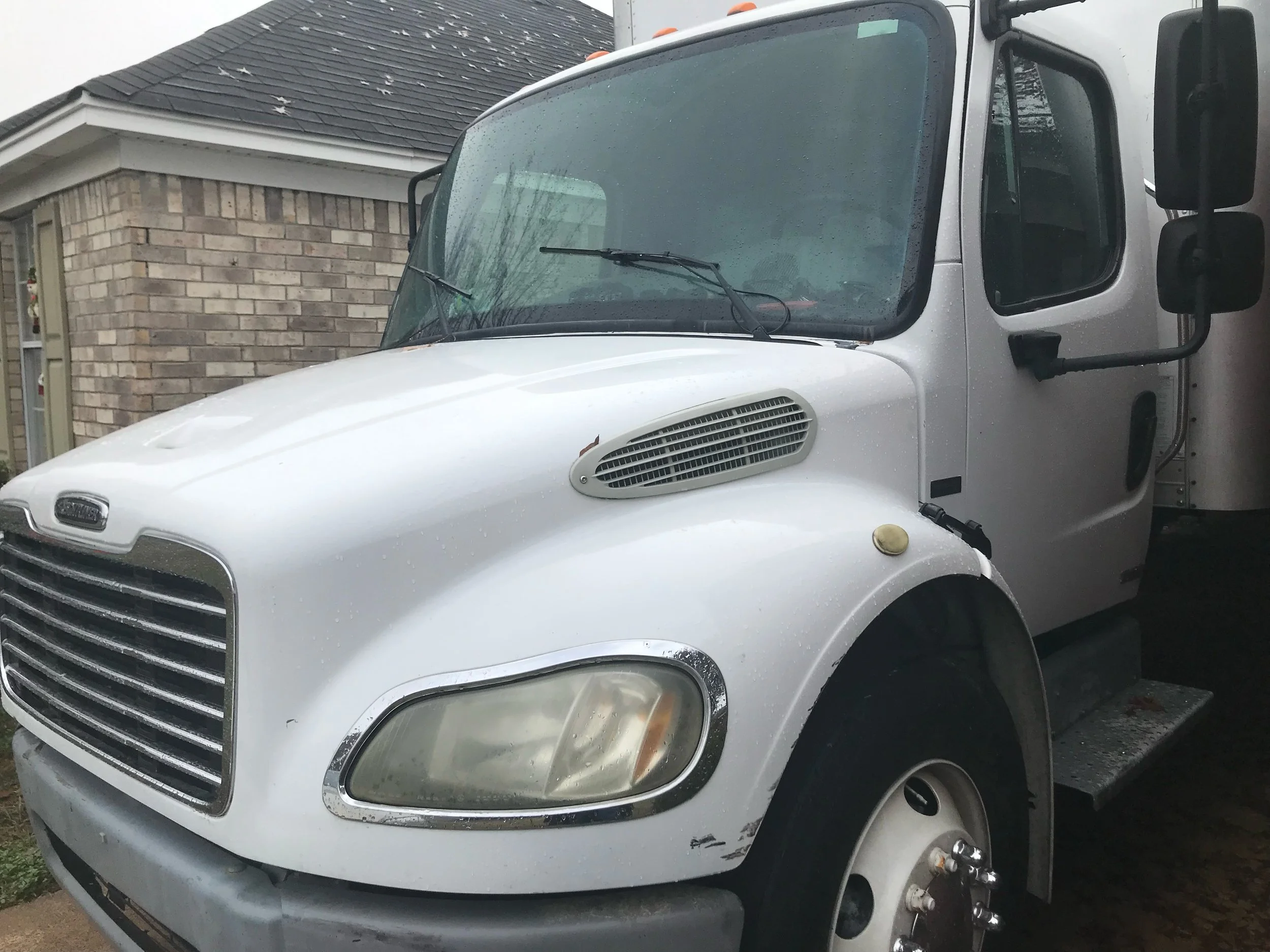 White box truck parked outside a house, with visible water droplets on the windshield and a dent on the front left side.