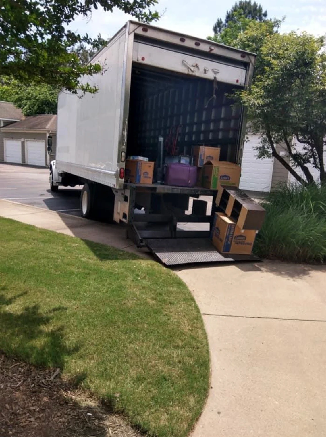 A moving truck with its back door open, filled with boxes, parked on a driveway next to a grassy area and a sidewalk.