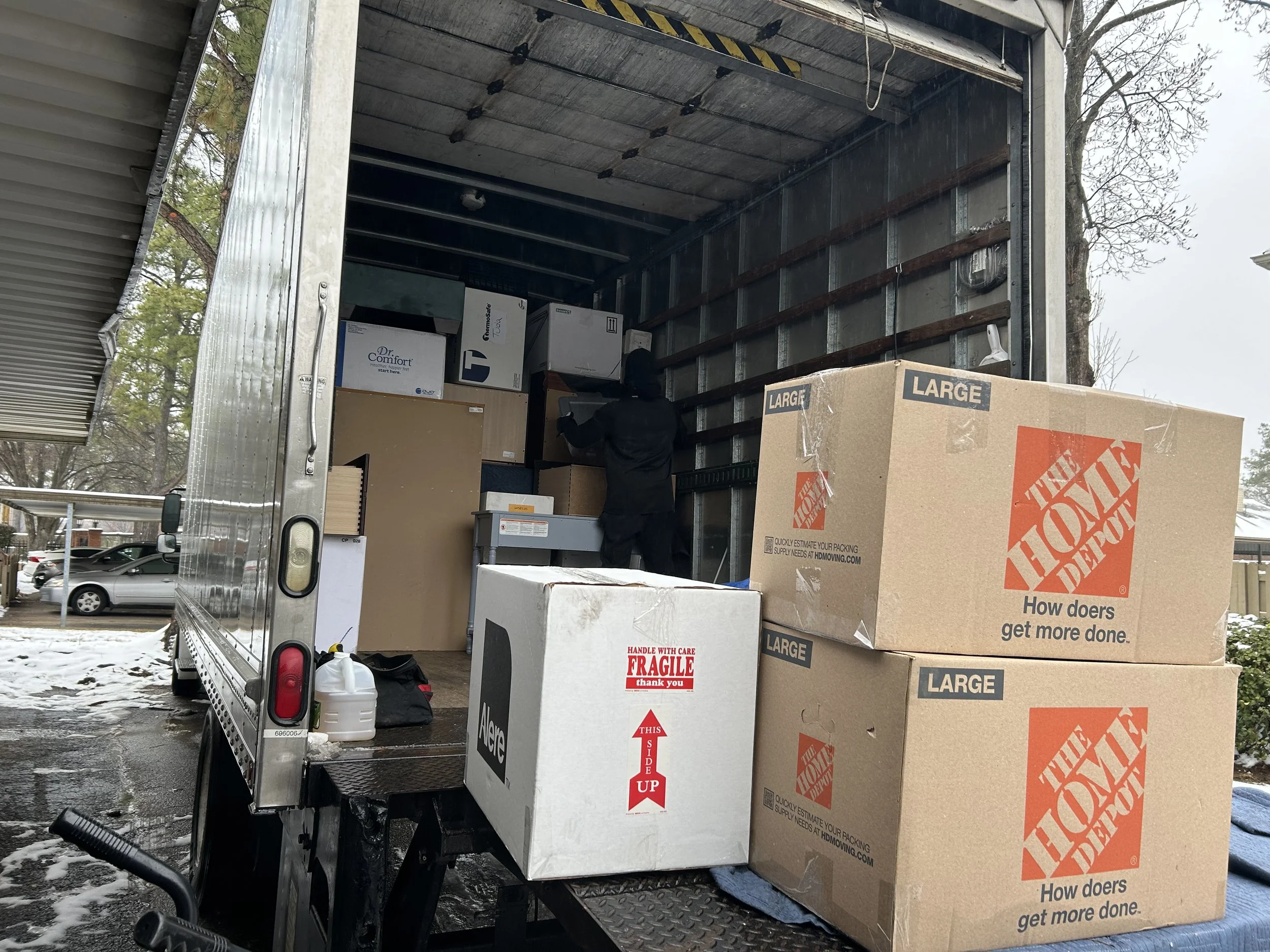 Open moving truck with cardboard boxes inside and on the ground nearby, parked outdoors on a snowy and wet day.