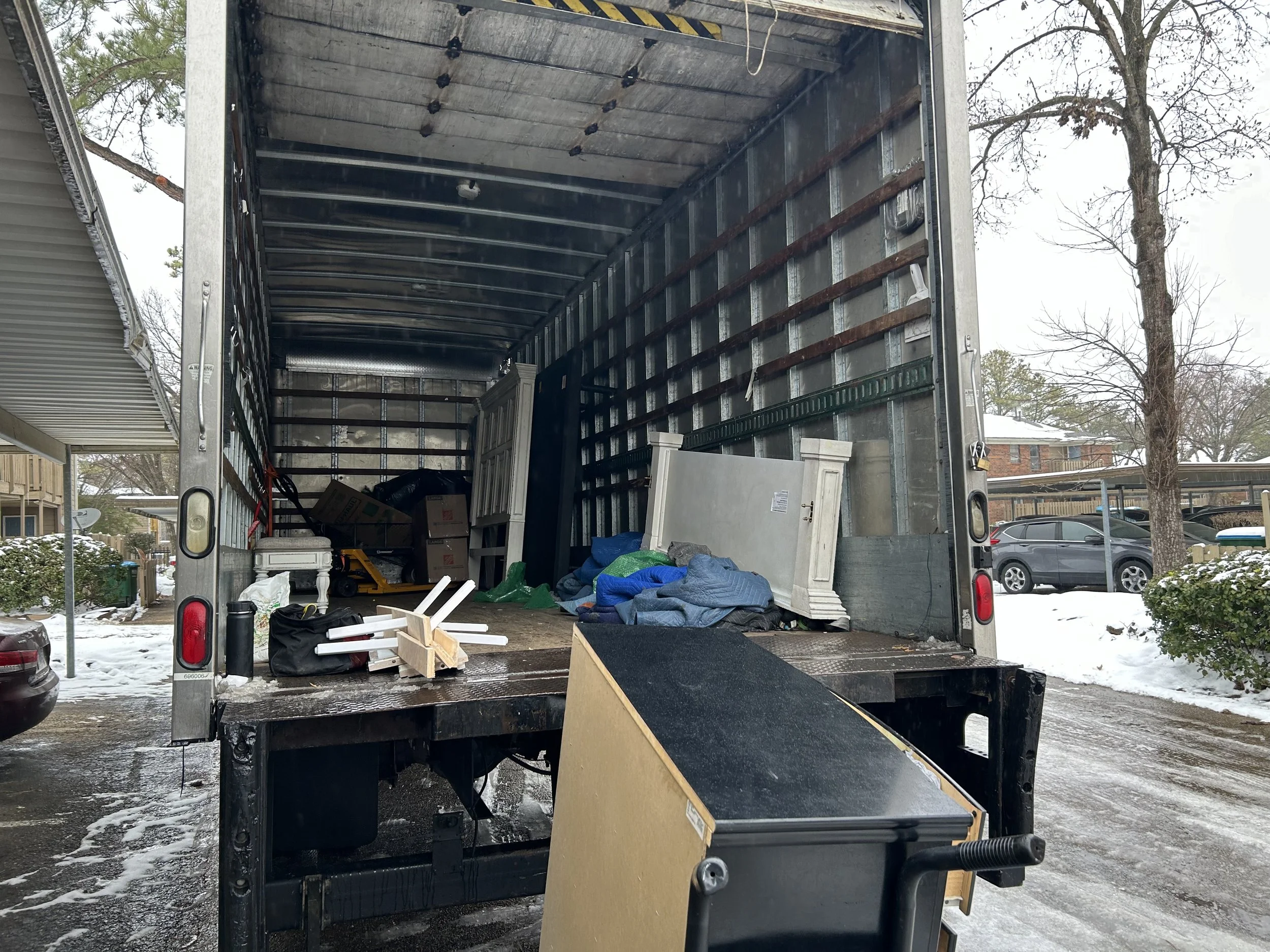 The back of an open moving truck parked in a residential area during winter, with various household items, boxes, furniture, and bags inside, and some items scattered outside the truck.