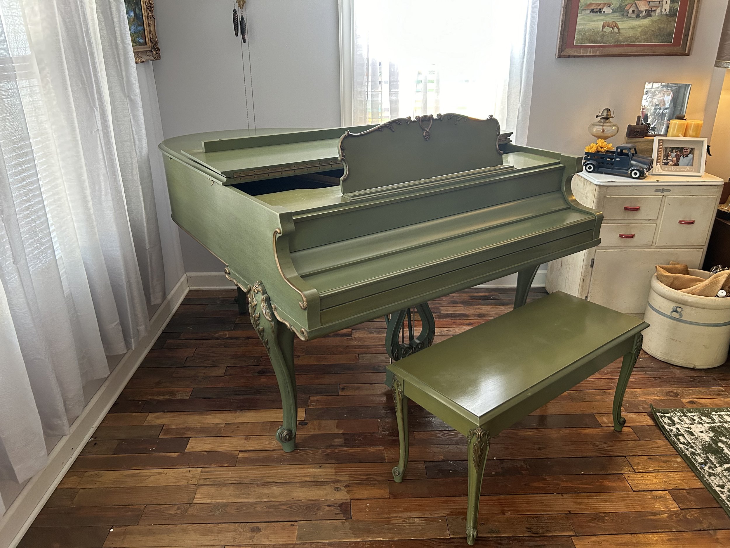A vintage green piano with ornate gold trim, a matching piano bench, and various decorative items on a white dresser, near a window with sheer curtains.