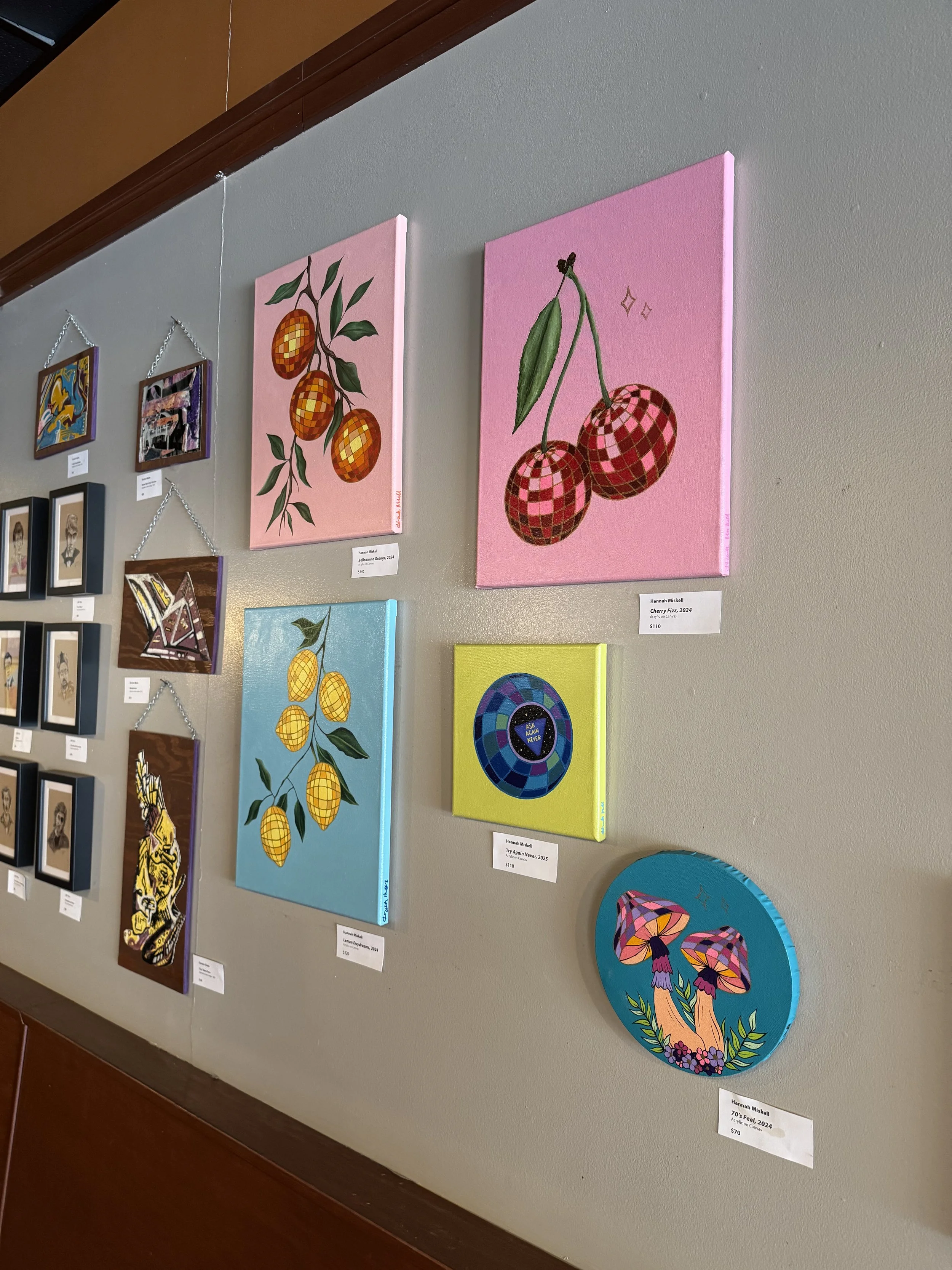 Art display with various colorful paintings, including pink backgrounds with stylized cherries and other vibrant designs, hanging on a gray wall.
