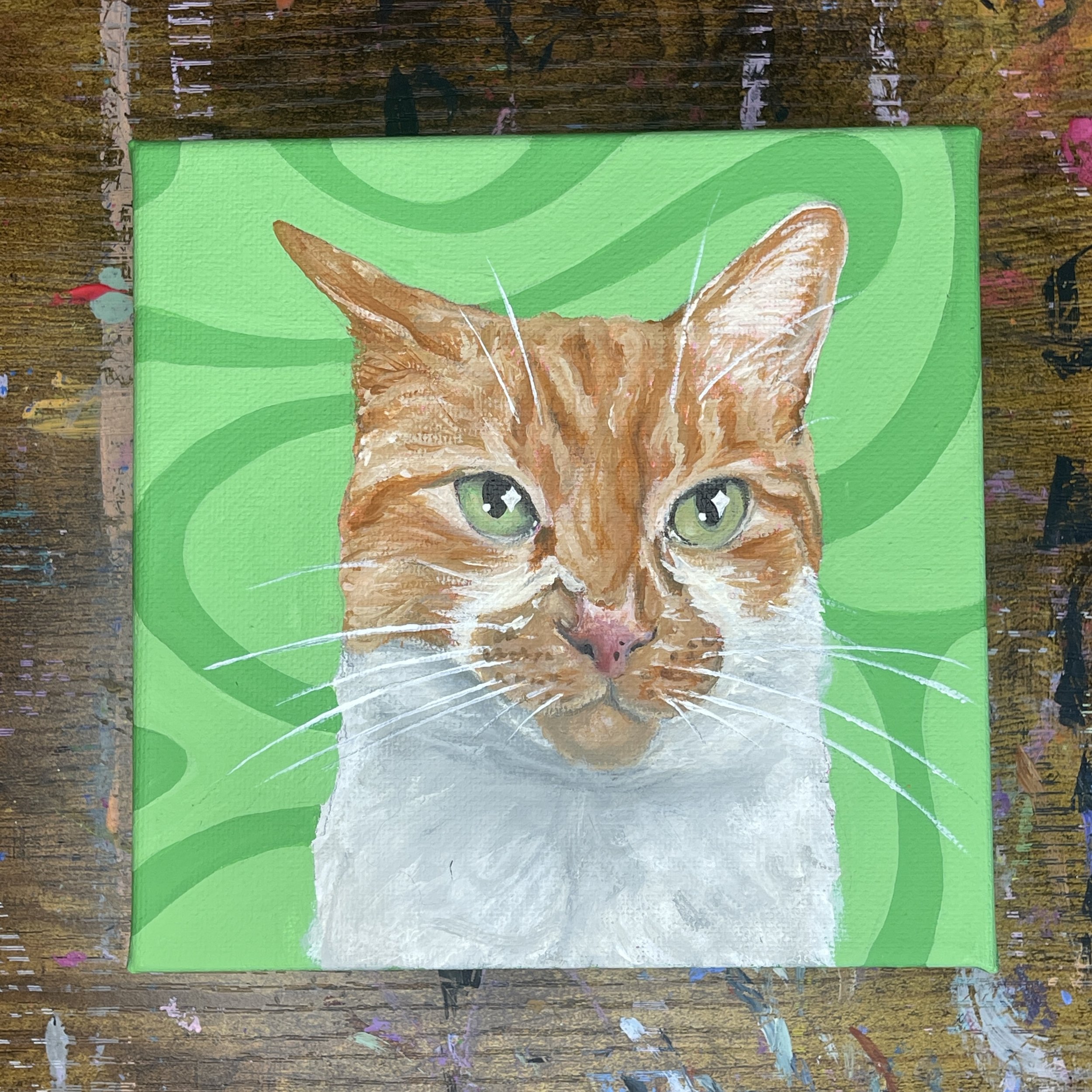 Painting of a cat's face with green eyes on a bright green background, placed on a wooden surface.