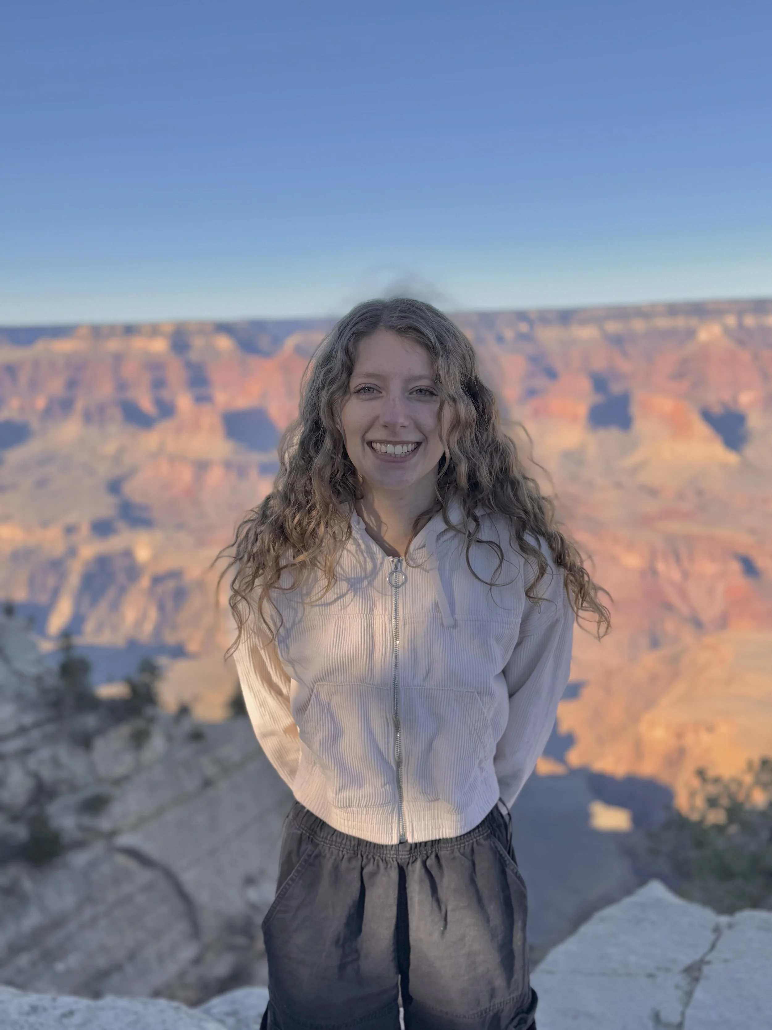 A young woman with curly hair smiling at the camera at the Grand Canyon during sunset or sunrise, with a clear blue sky in the background.