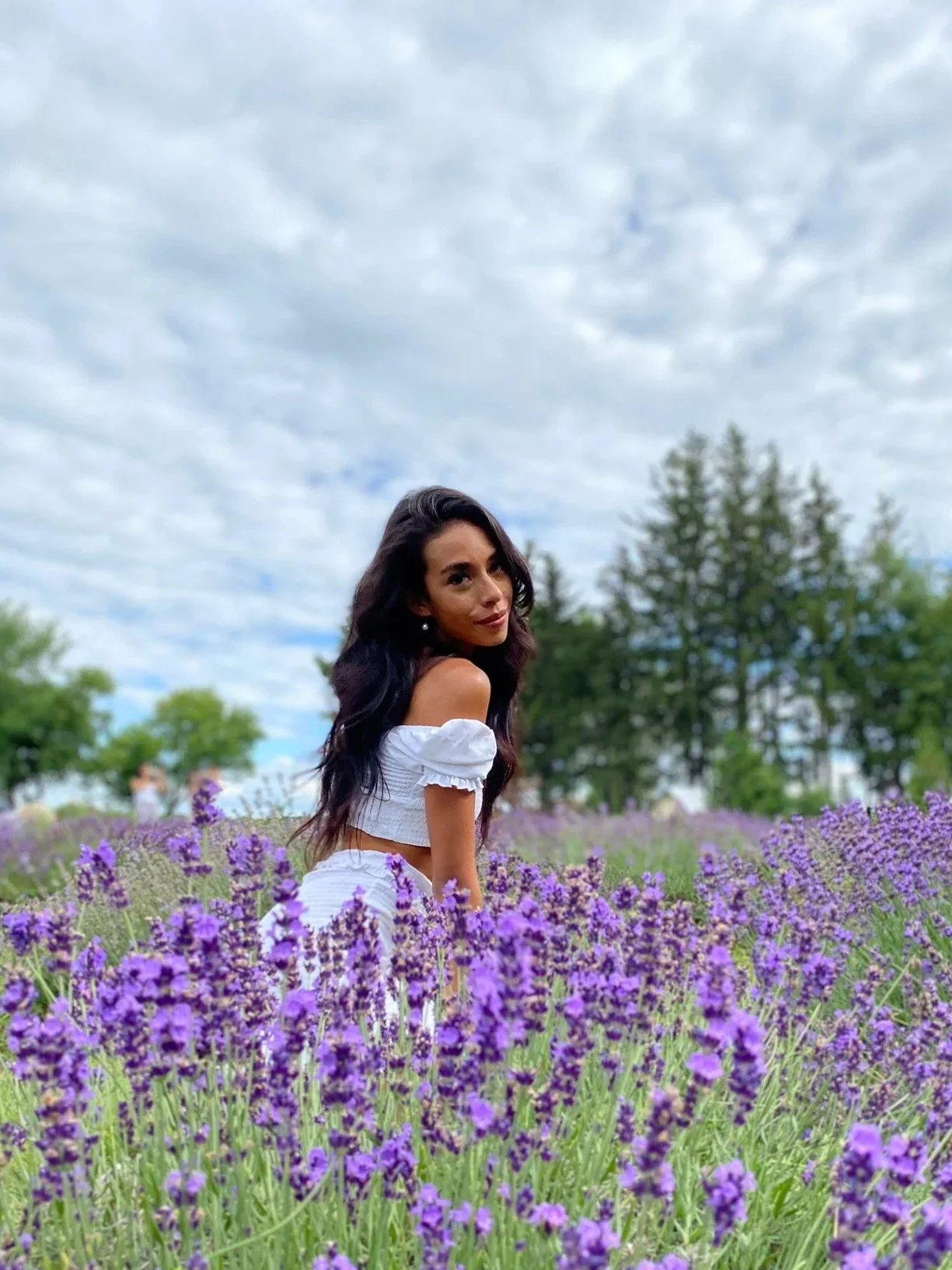 A woman in a white off-shoulder top and skirt standing among purple lavender flowers in a field with trees and cloudy sky in the background.
