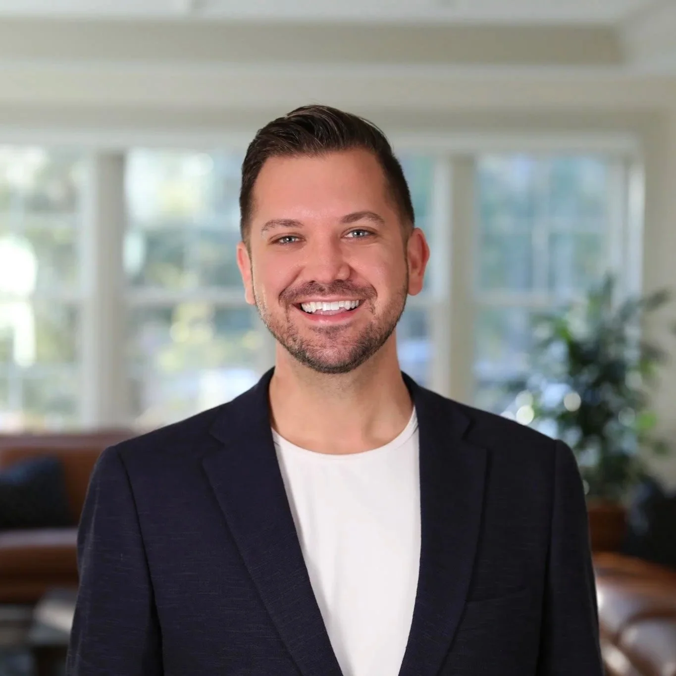 A man with short dark hair, a beard, and a big smile standing in a well-lit living room with large windows in the background, wearing a dark blazer and a white shirt.