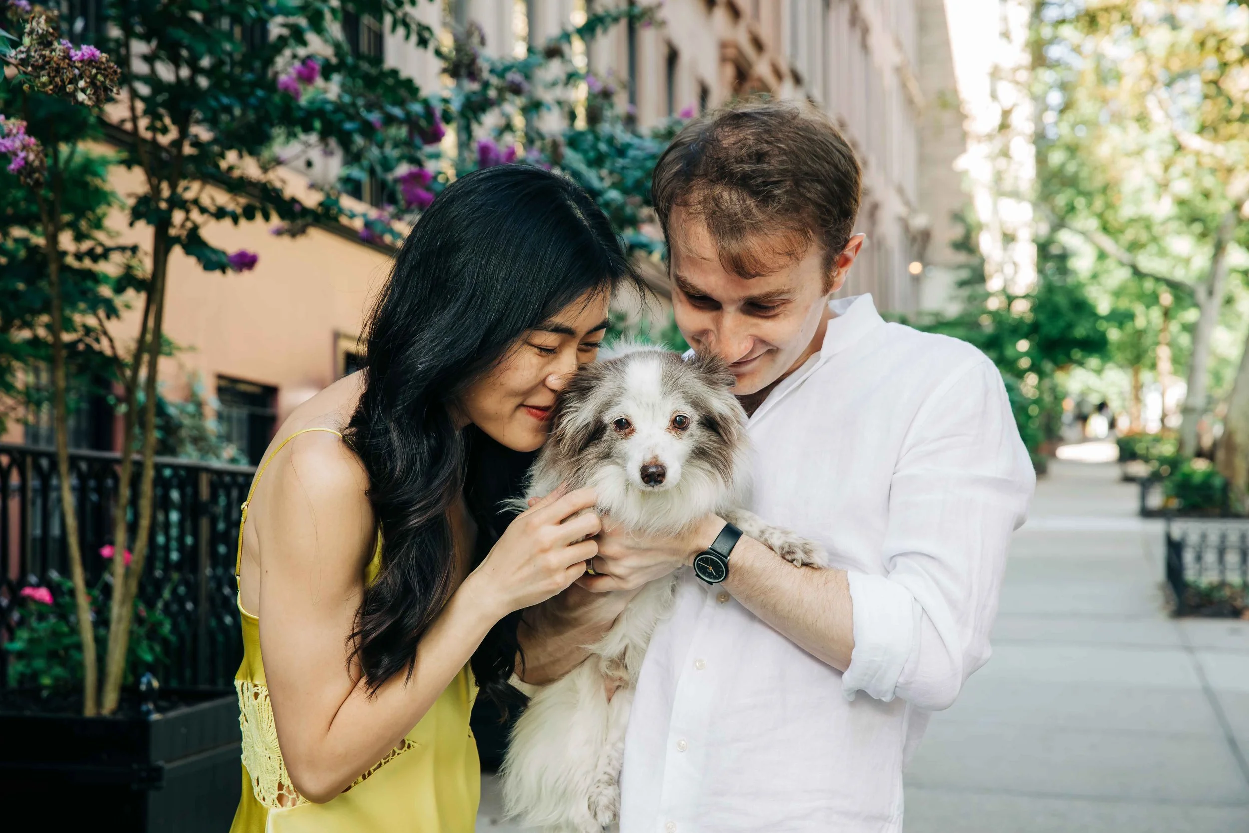 A couple holding a dog outdoors on a city sidewalk, with trees and flowers in the background.