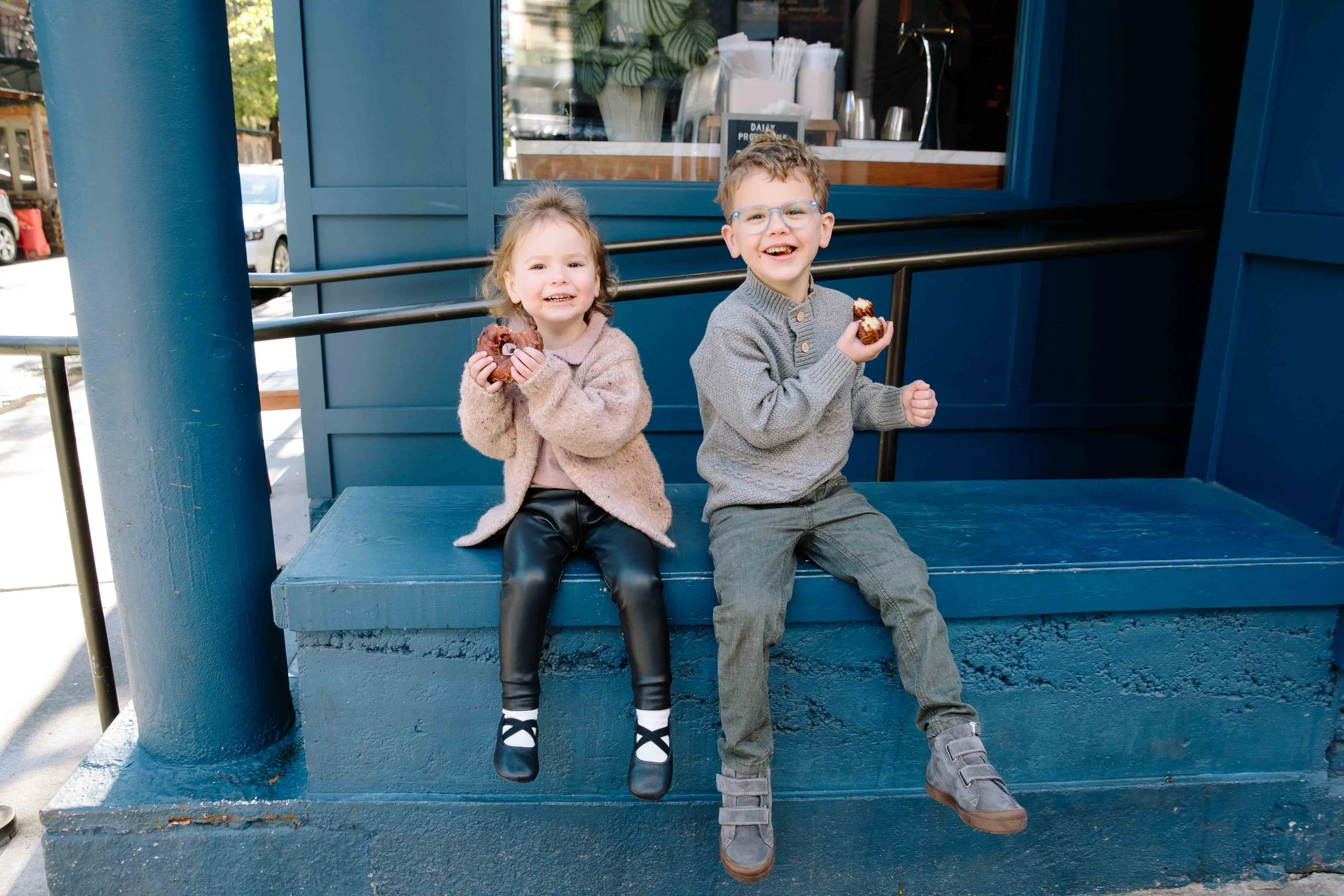 Two children, a girl and a boy, sitting on a blue bench outside a building, eating donuts and smiling at the camera.