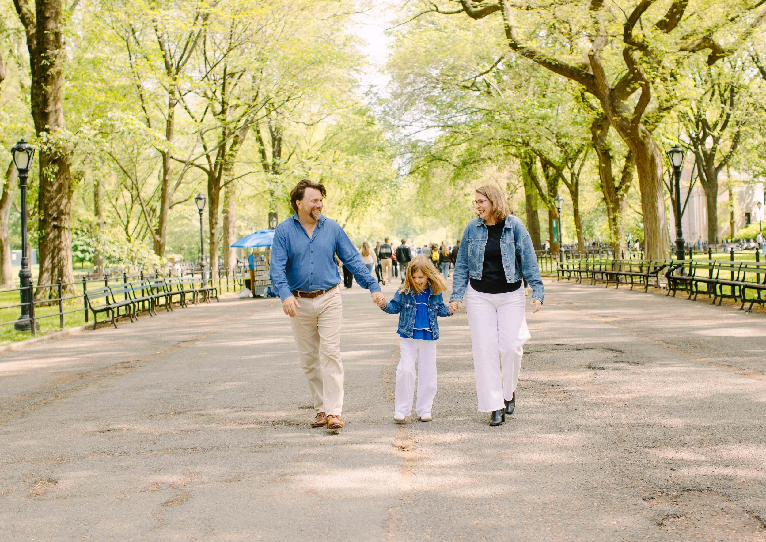 A family of three, a man, a woman, and a girl, walking hand in hand in a park with green trees and benches, during spring.