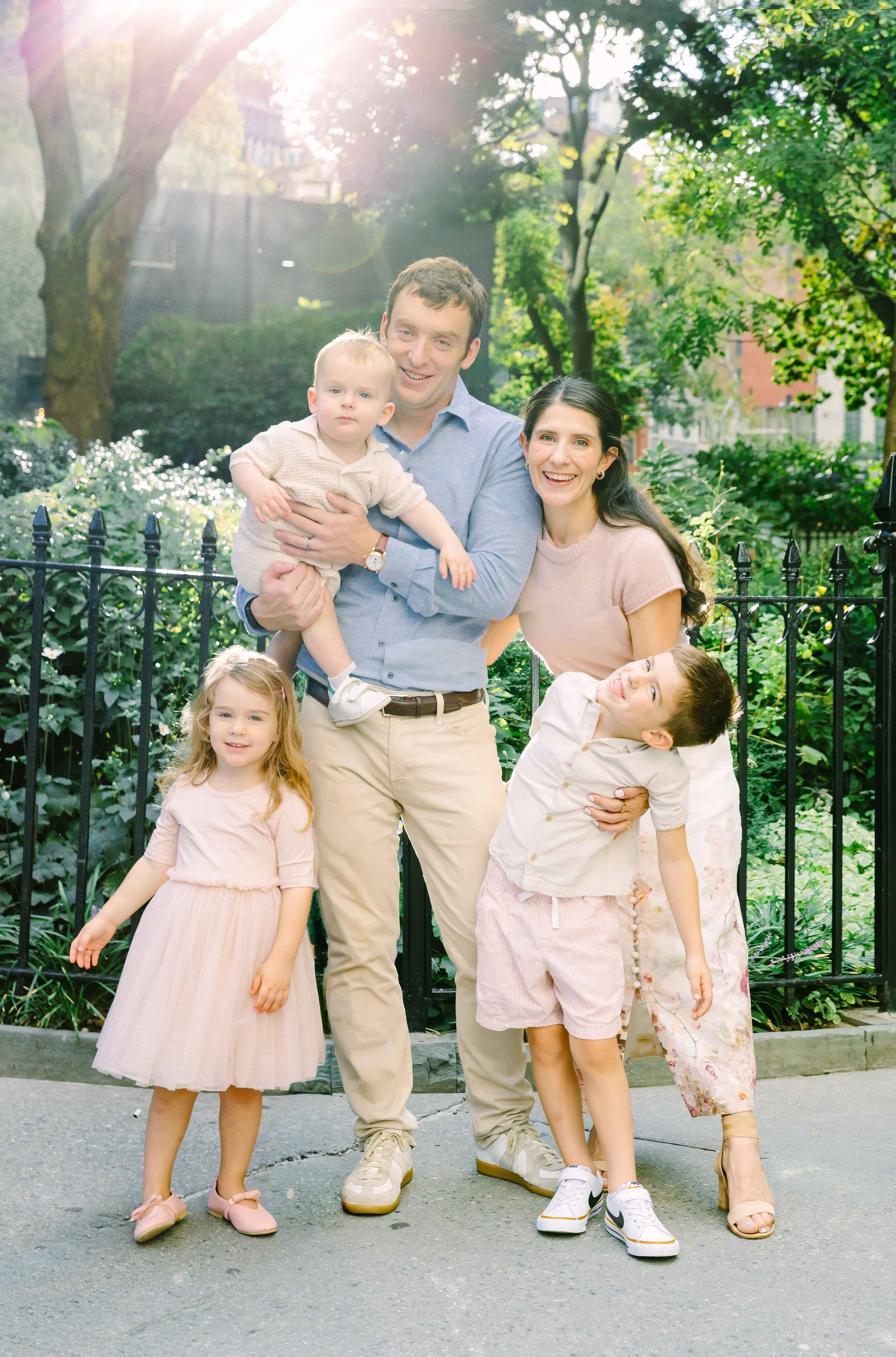 A family of six posing outdoors in a park with trees and a black iron fence. The father is holding a toddler son, and the mother and four children are standing around them. The sunlight is shining through the trees.