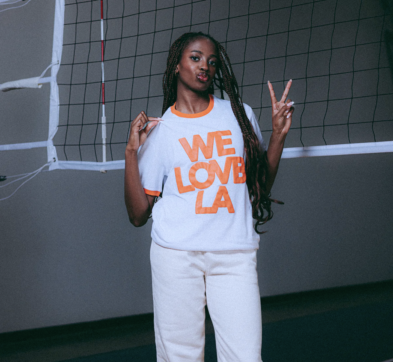 A woman with long braided hair standing in front of a volleyball net, wearing a white t-shirt with orange lettering that says 'WE LOVE LA', and white pants, making peace signs with both hands.
