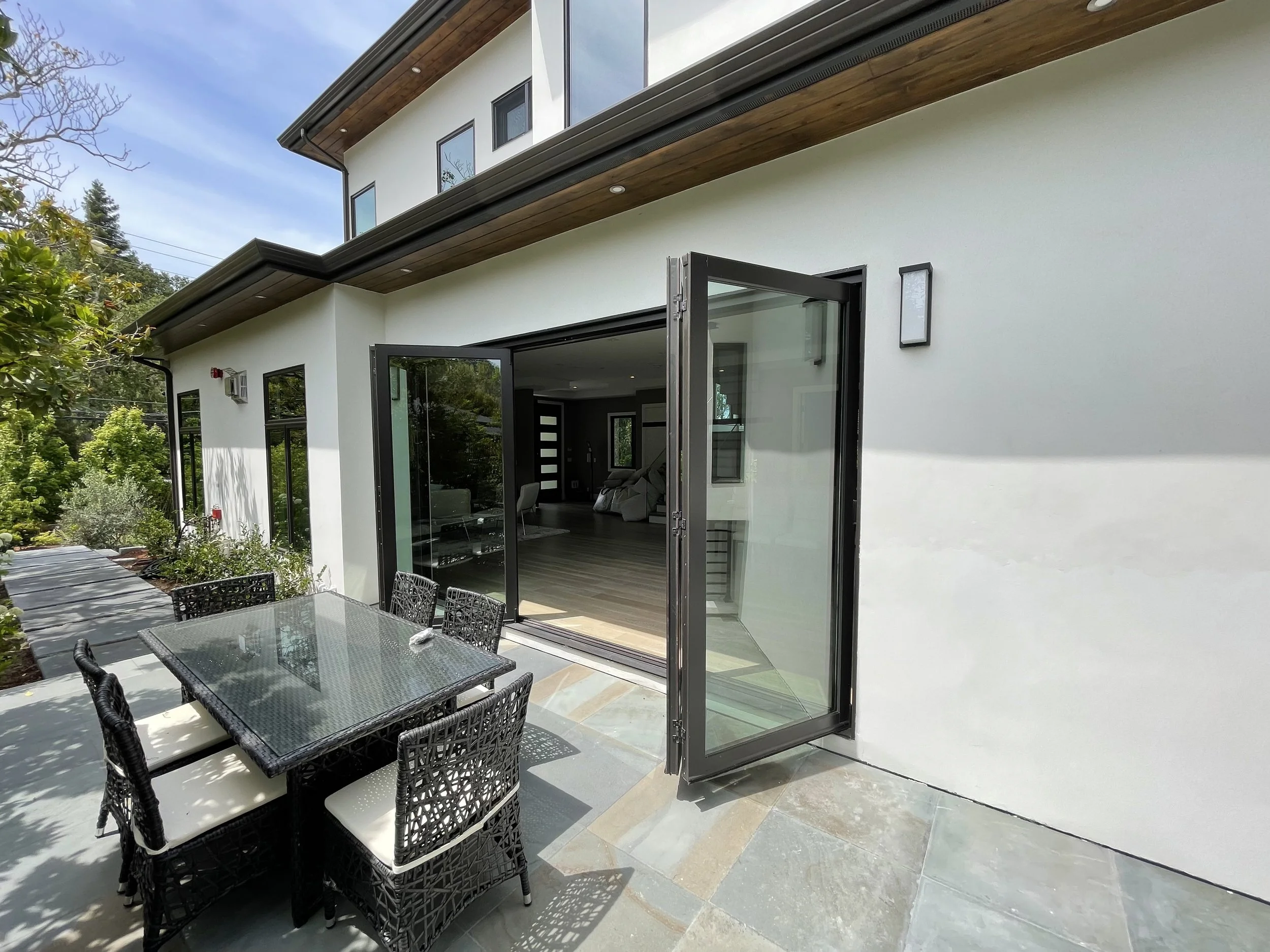 Modern house with large glass doors opening to an outdoor patio with a black wicker dining set, surrounded by greenery and trees, under a blue sky.