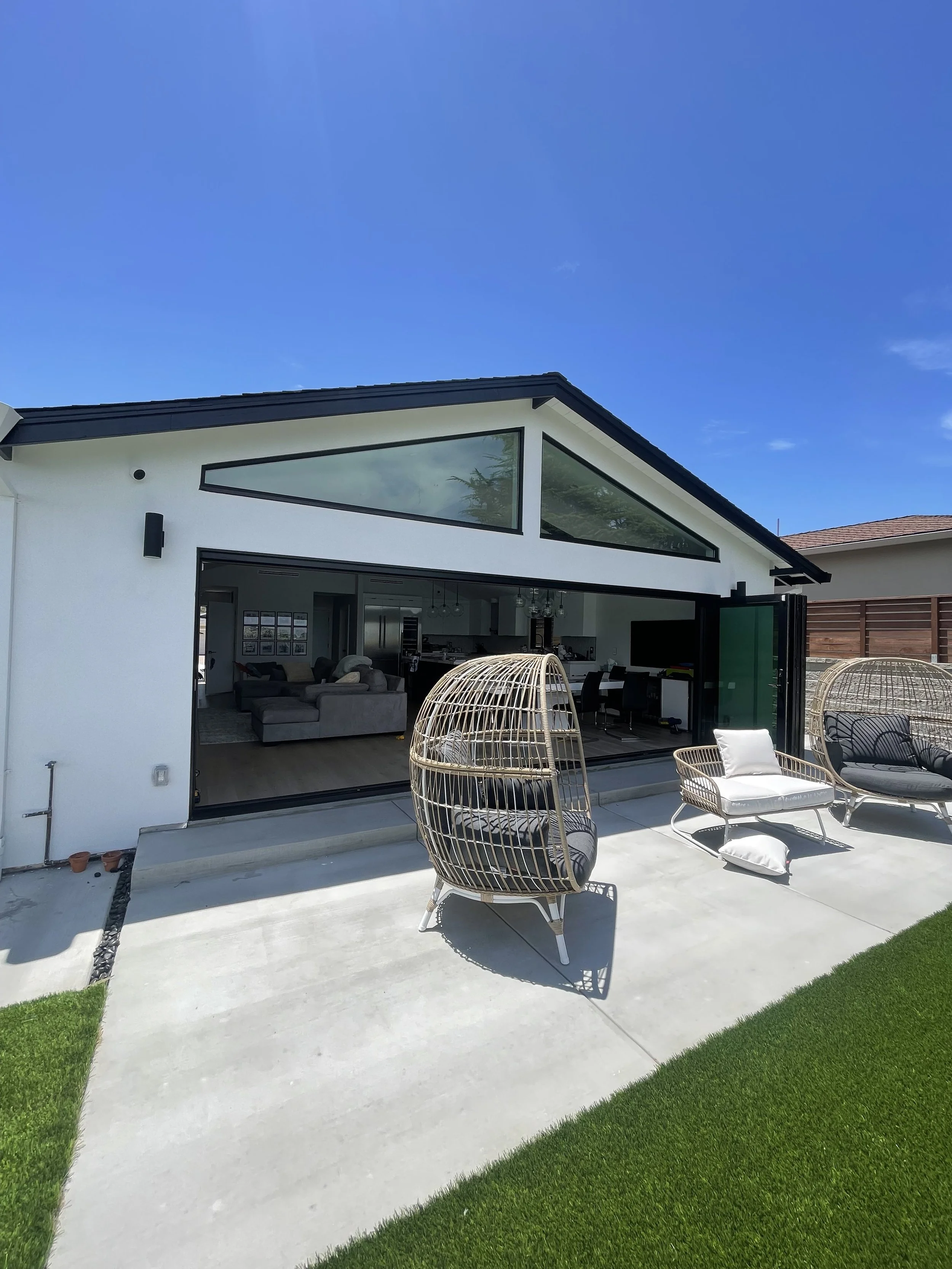 Modern house with large glass windows open to outdoor patio with chairs and a small grassy area.