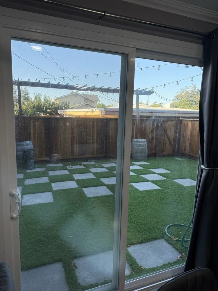 View of a backyard through a sliding glass door, featuring artificial grass, a checkered pattern of concrete pavers, string lights overhead, a wooden fence, and a house in the background.