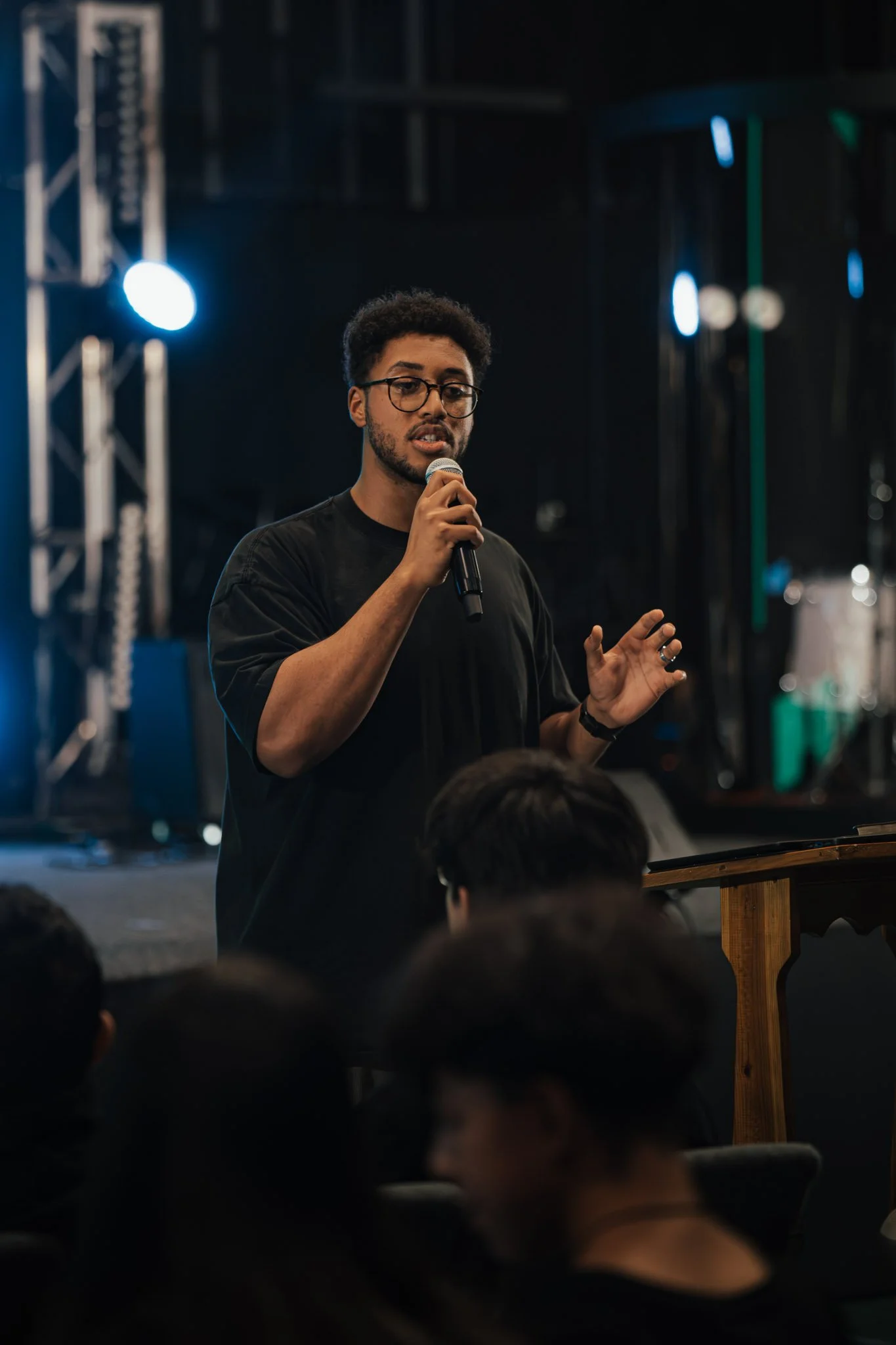 A man with short curly hair, glasses, and wearing a black shirt, speaking into a microphone during a presentation or speech at an indoor event.