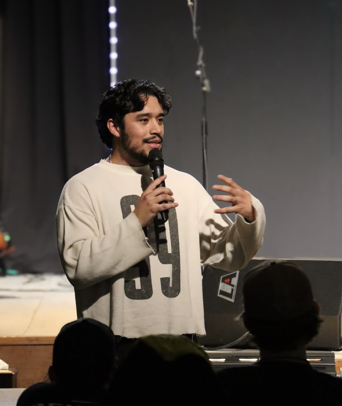 A young man with dark hair and a beard, holding a microphone, speaking on stage in front of an audience, wearing a white sweatshirt with black numbers.