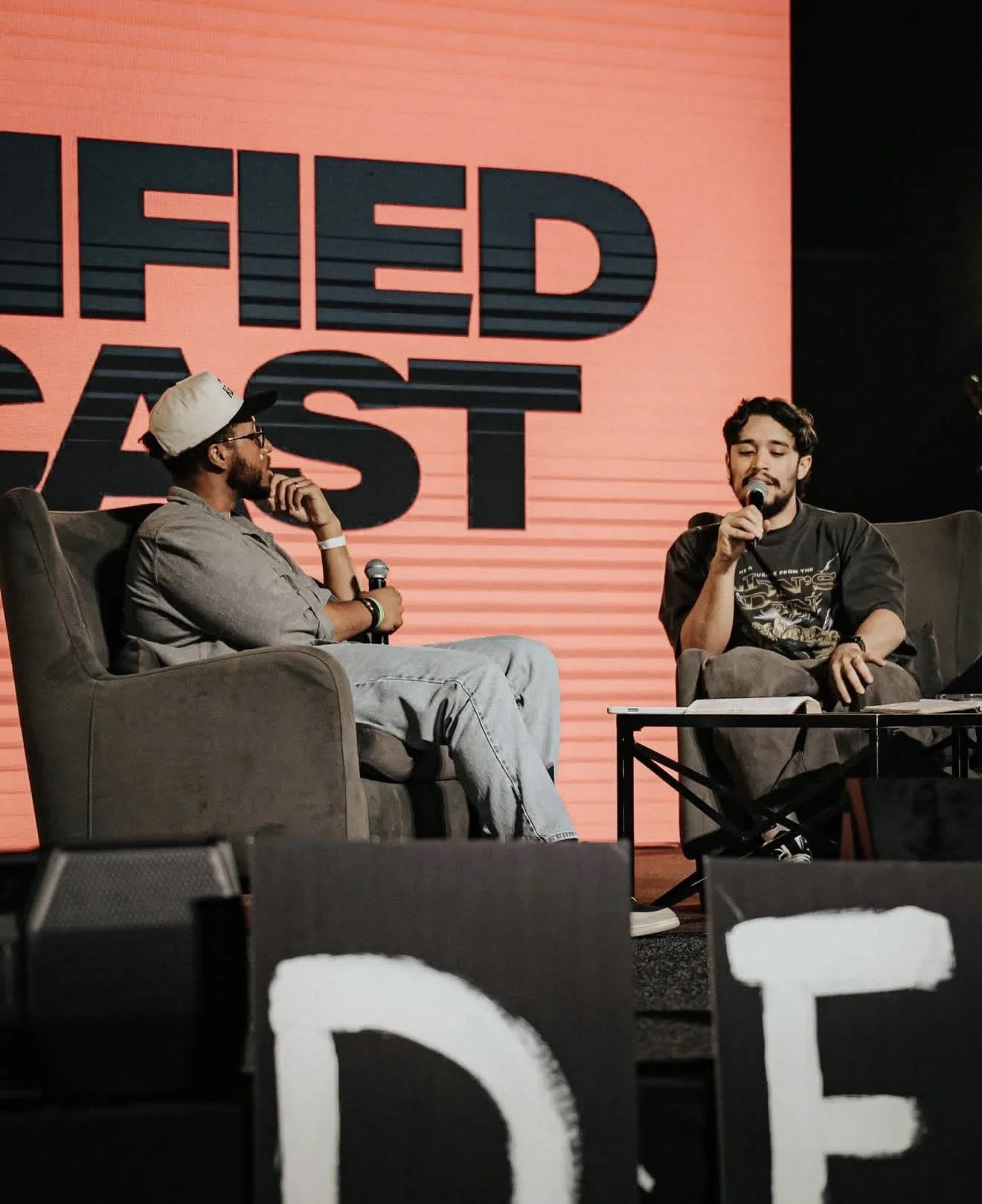 Two men are sitting on stage in chairs, holding microphones, engaging in a discussion during an event with a red and black background that reads 'THE LAST'.