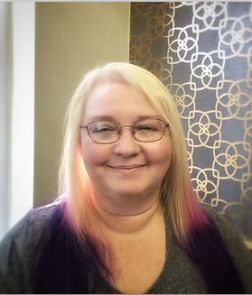 A woman with blonde hair, glasses, and a smile, standing indoors in front of a patterned wall.