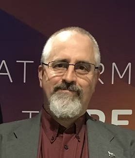 A middle-aged man with short gray hair and a beard, wearing glasses and a gray blazer over a maroon shirt, standing in front of a purple background.