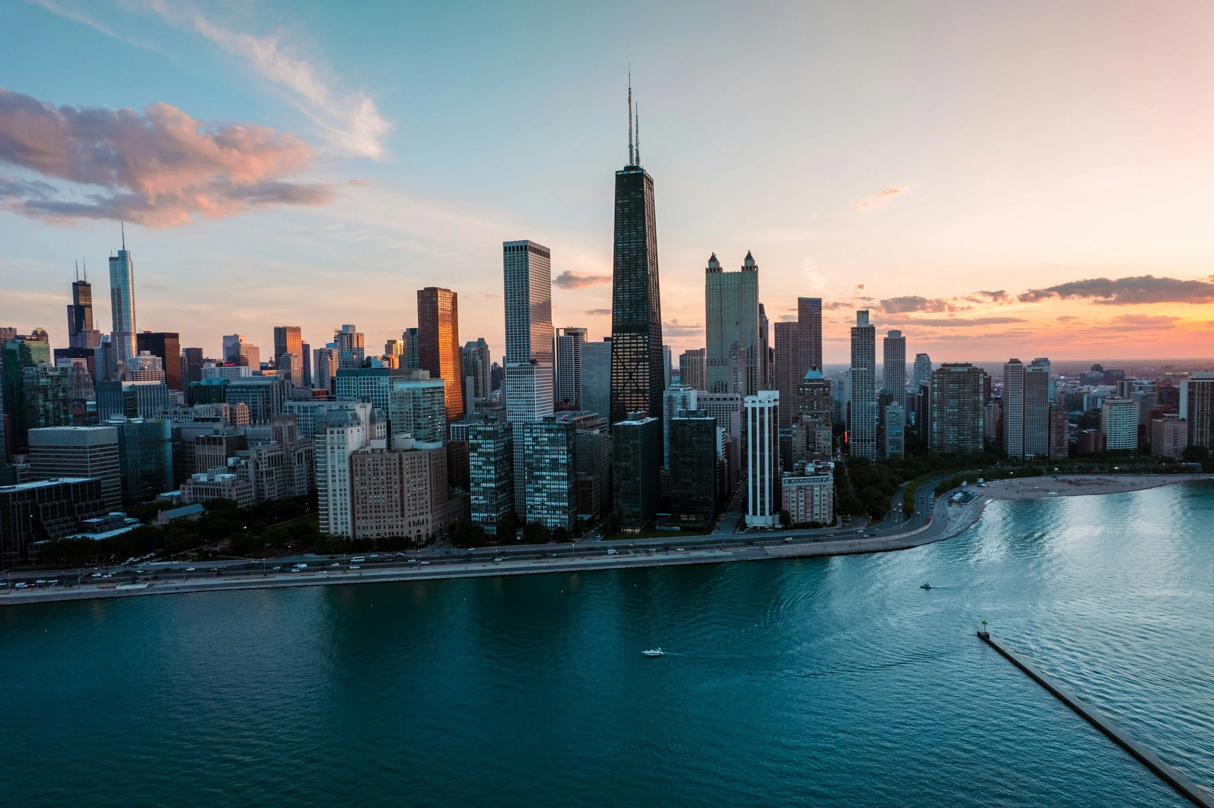 Aerial view of Chicago skyline at sunset, featuring tall buildings including the Willis Tower and John Hancock Center, with a body of water in the foreground and a partly cloudy sky.