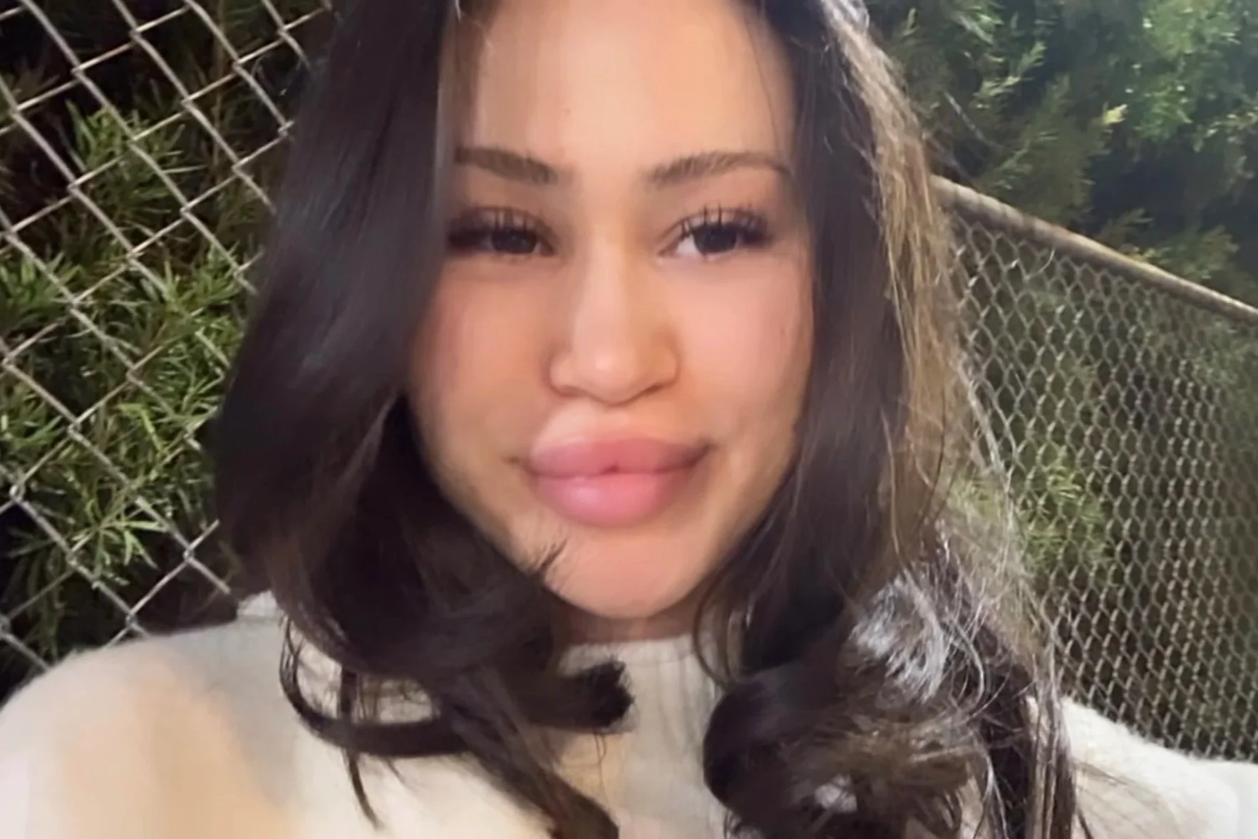 Close-up of a young woman with long, dark hair, smiling gently, standing outdoors near a chain-link fence with green foliage in the background.