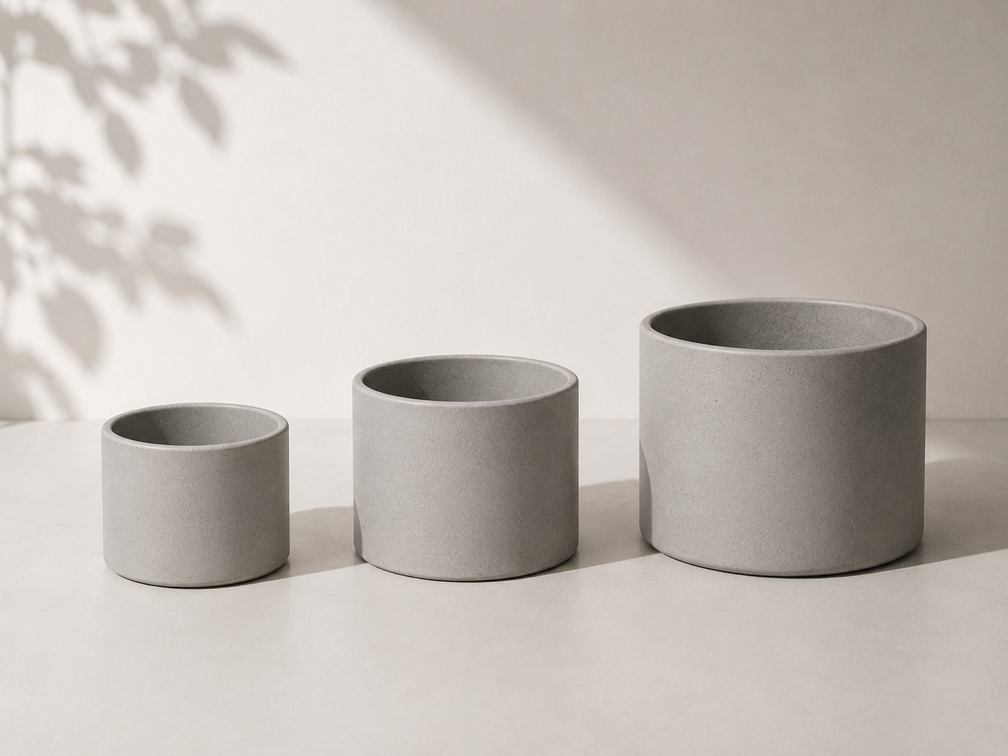 Três vasos de cerâmica cinza em tamanhos diferentes, alinhados em uma superfície de tampo claro com sombra suave ao fundo.