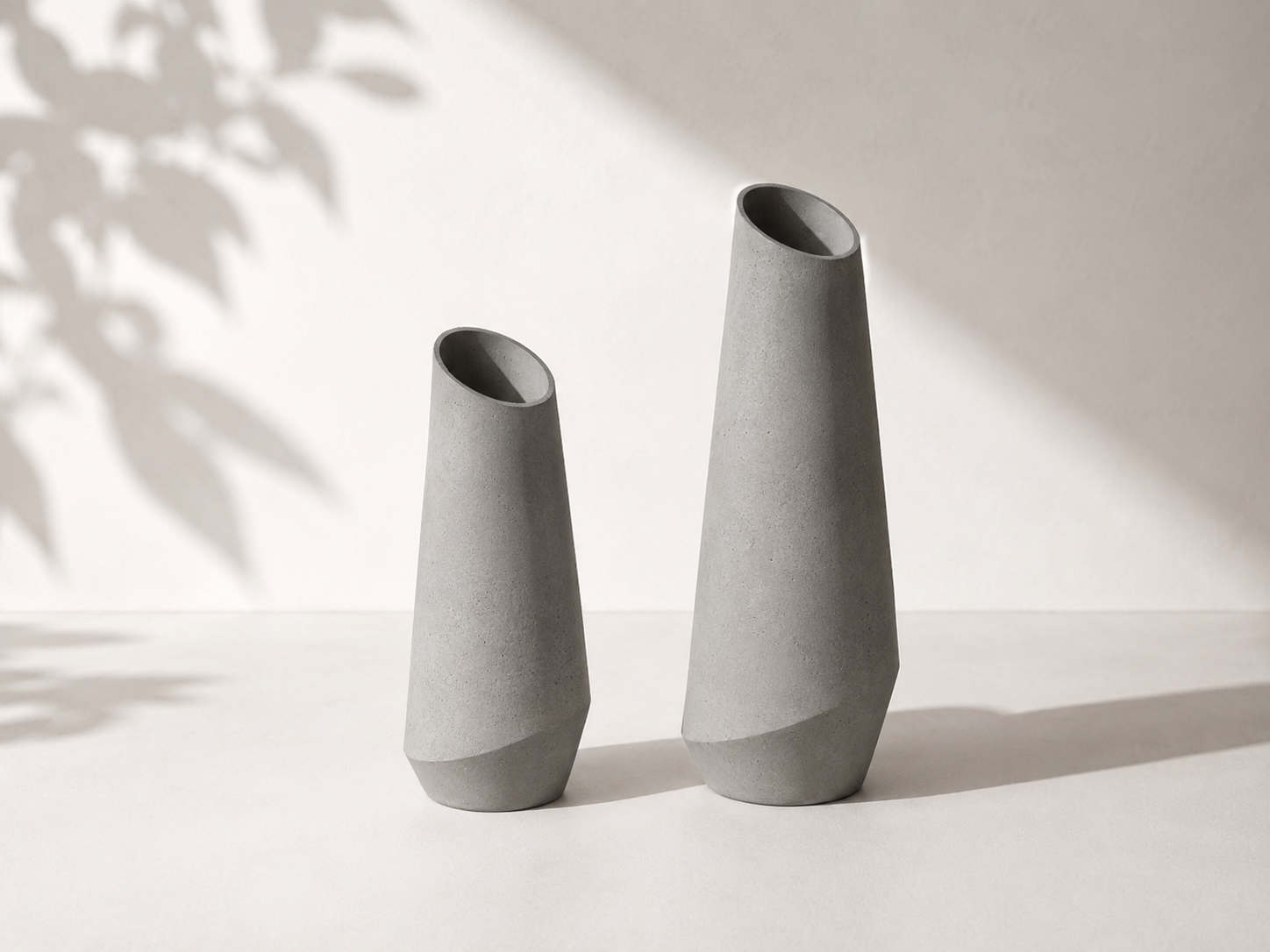 Dois vasos de concreto cinza de formatos diferentes, com sombra projetada na superfície branca ao fundo, e uma sombra de folha de árvore no lado esquerdo.