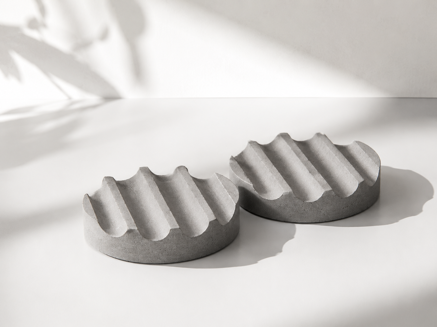 Dois objetos de concreto cinza com formato de meia-lua, com ranhuras retas na parte superior, colocados sobre uma superfície branca com sombra e iluminação suave.