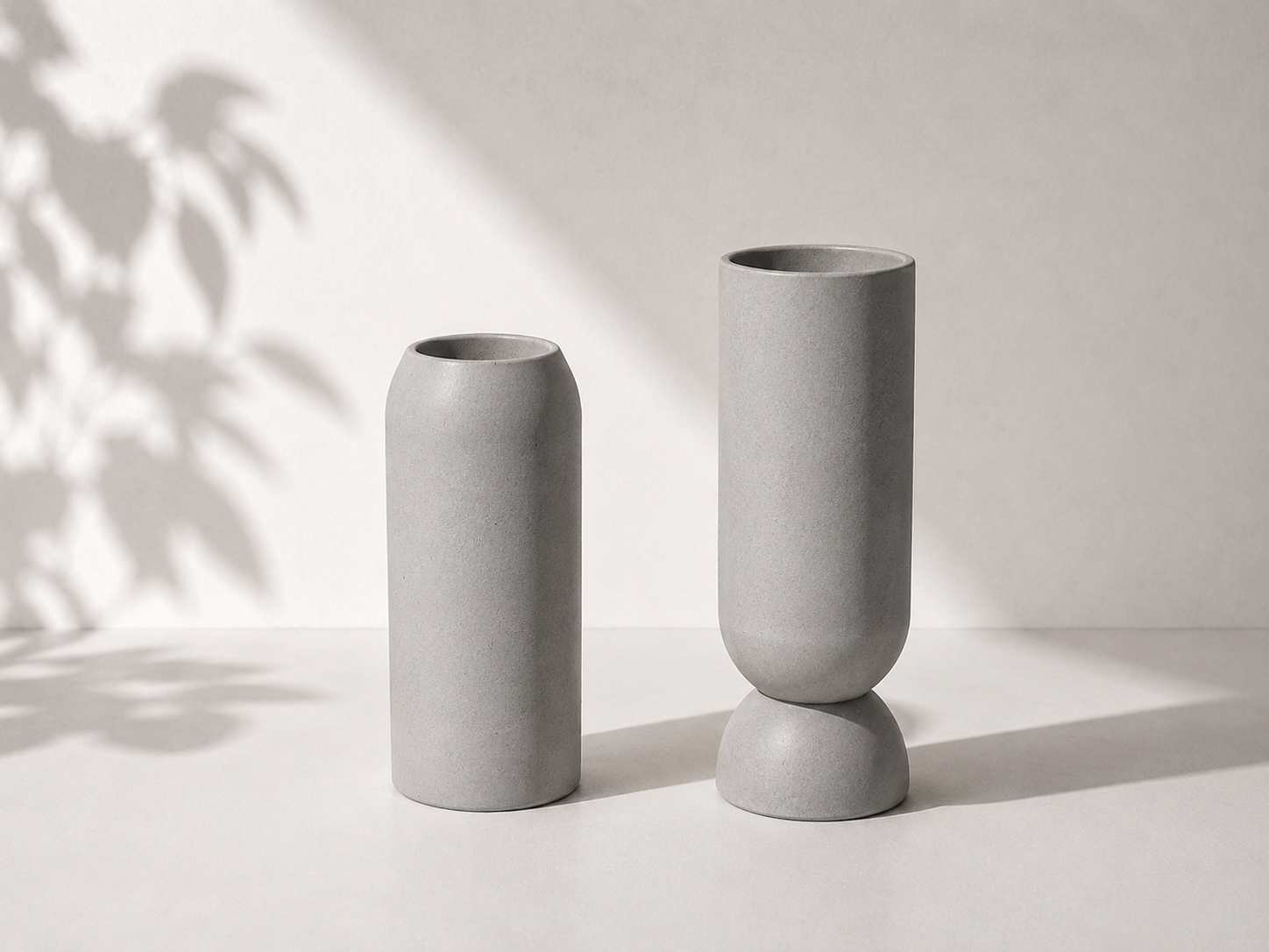 Dois vasos cilíndricos de concreto de diferentes alturas, um apoiado em uma forma de pedra e o outro na superfície, com sombra de folhas na parede ao fundo.
