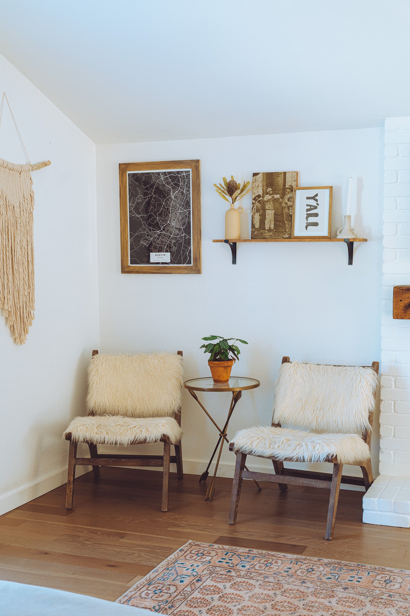 A cozy corner with two chairs with fur cushions, a small round table with a plant in a terracotta pot, a wall shelf with decorative items, and framed artwork on white walls, with a patterned rug on a wooden floor.