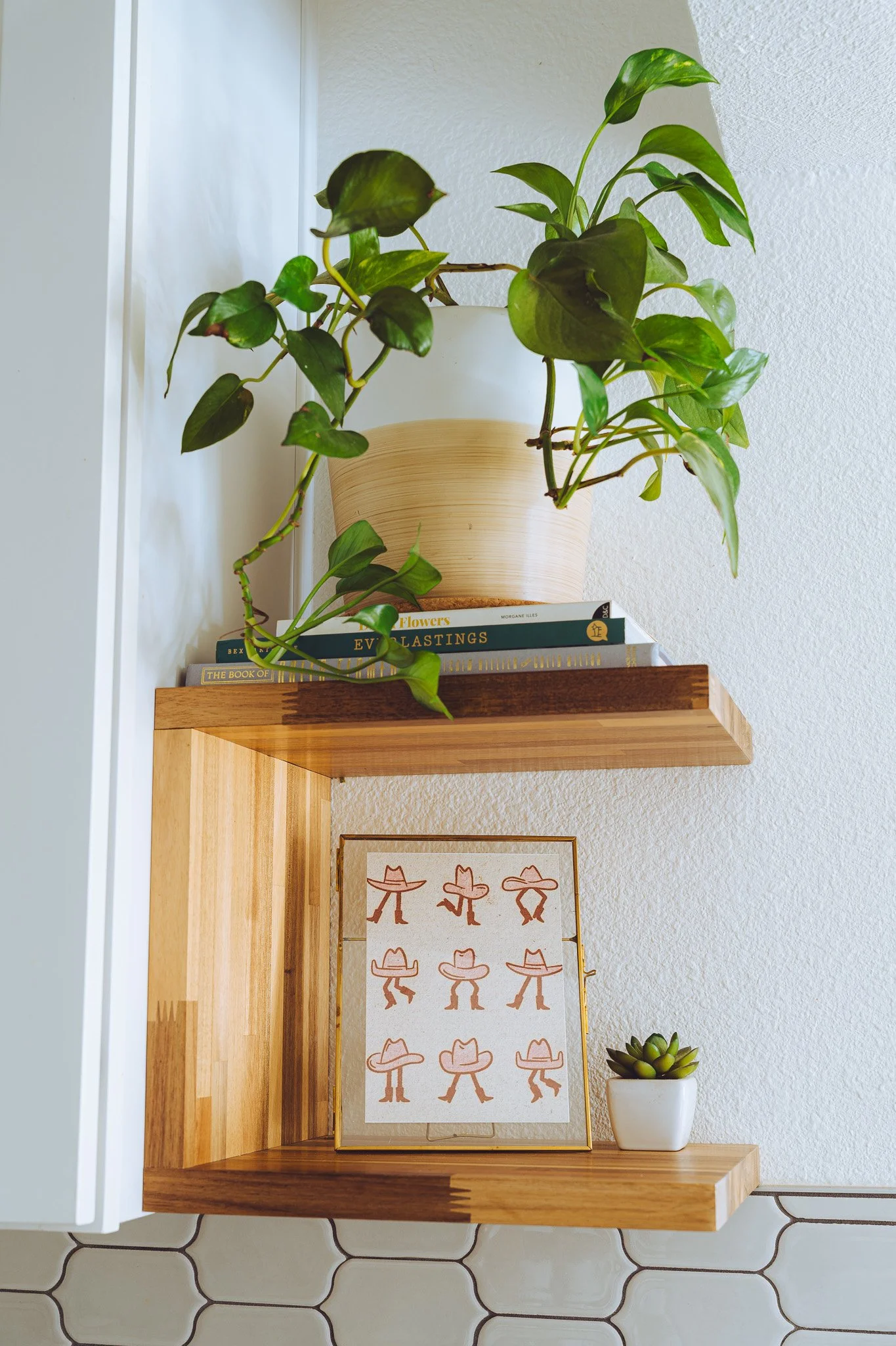 A wooden wall shelf with a potted plant on top, a framed picture of people wearing large hats, and a small succulent in a white pot. The background includes a textured white wall and hexagonal tile flooring.