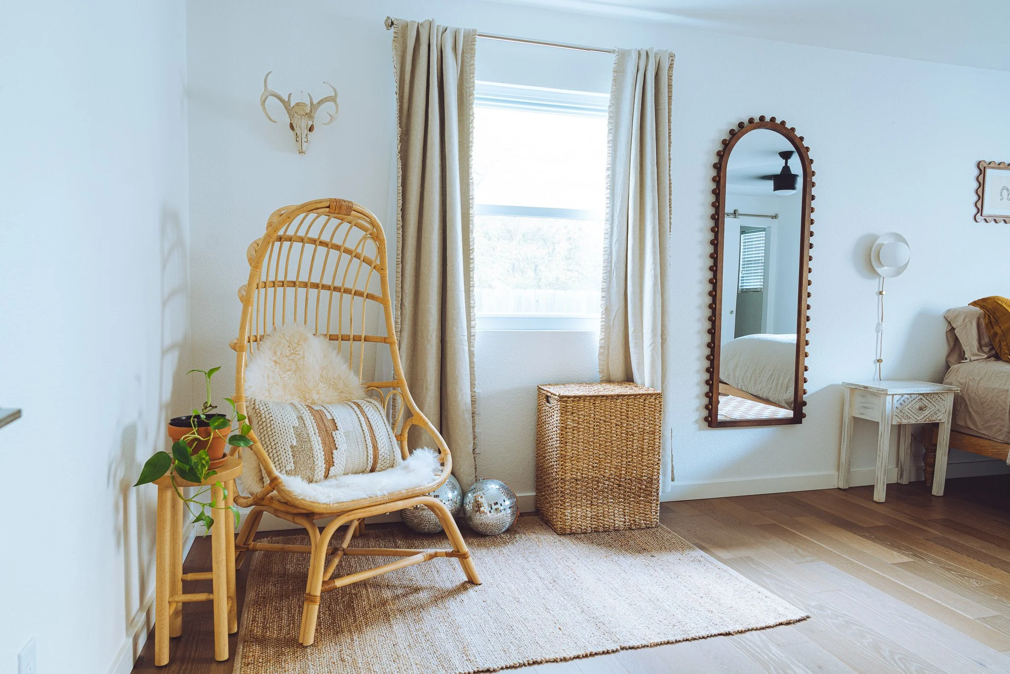 A cozy corner of a bedroom with a wicker chair, potted plant, curtains, a mirror, a small table, and decorative items like disco balls and a mounted animal skull on the wall.