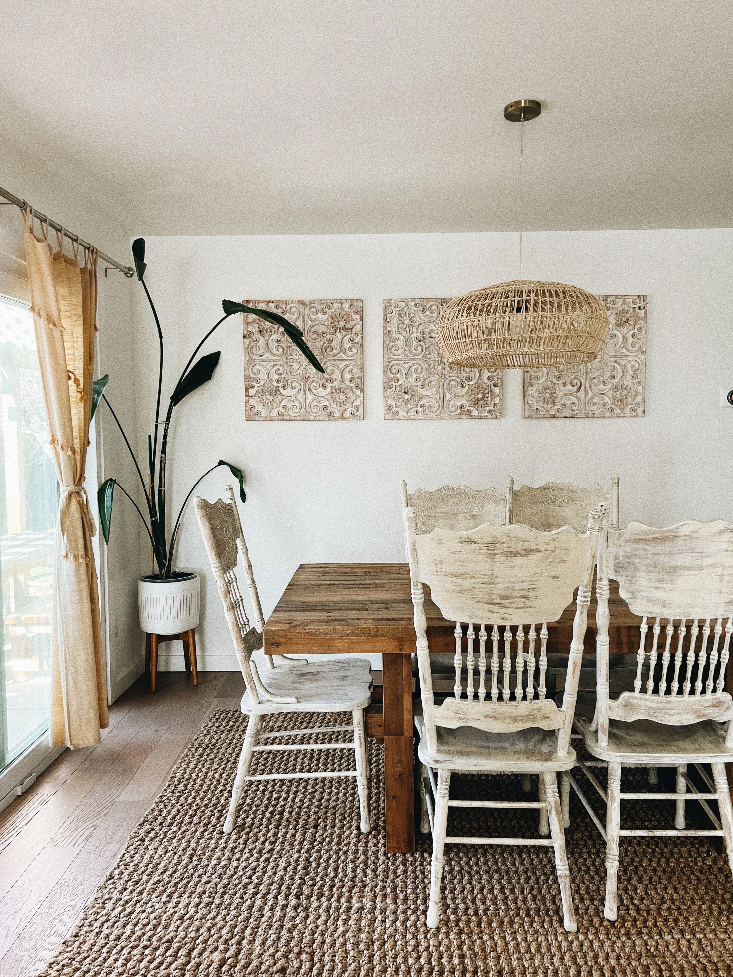 Dining room with wooden table, white distressed chairs, potted plant, woven basket pendant lamp, and textured wall art