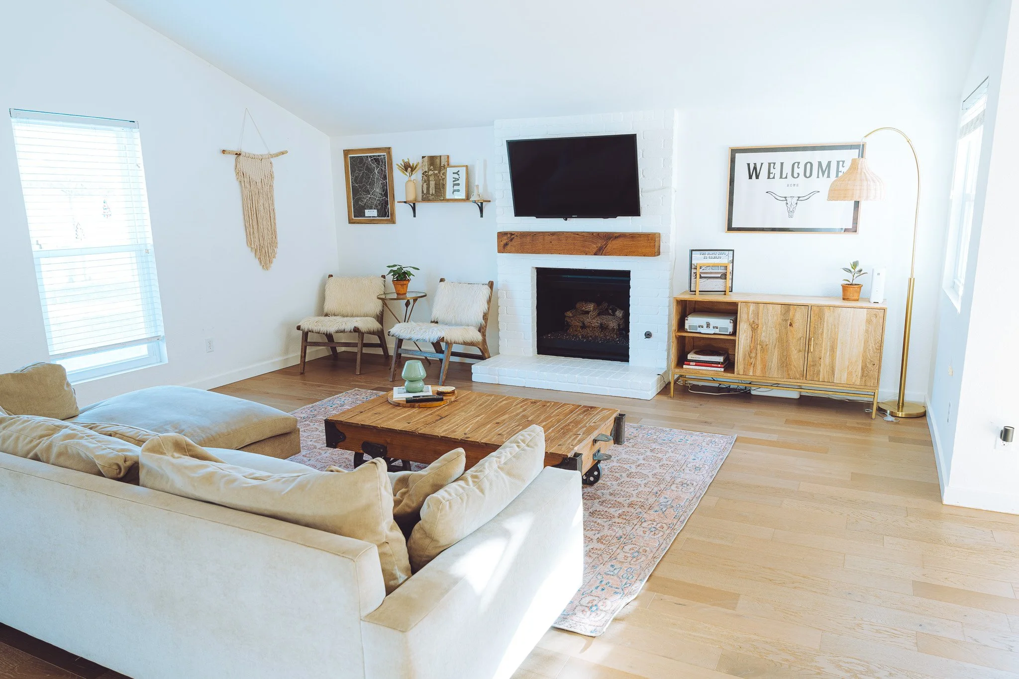 Bright living room with beige sofa, wooden coffee table, white brick fireplace, wall-mounted TV, and decor including framed artwork, plants, and a welcome sign.