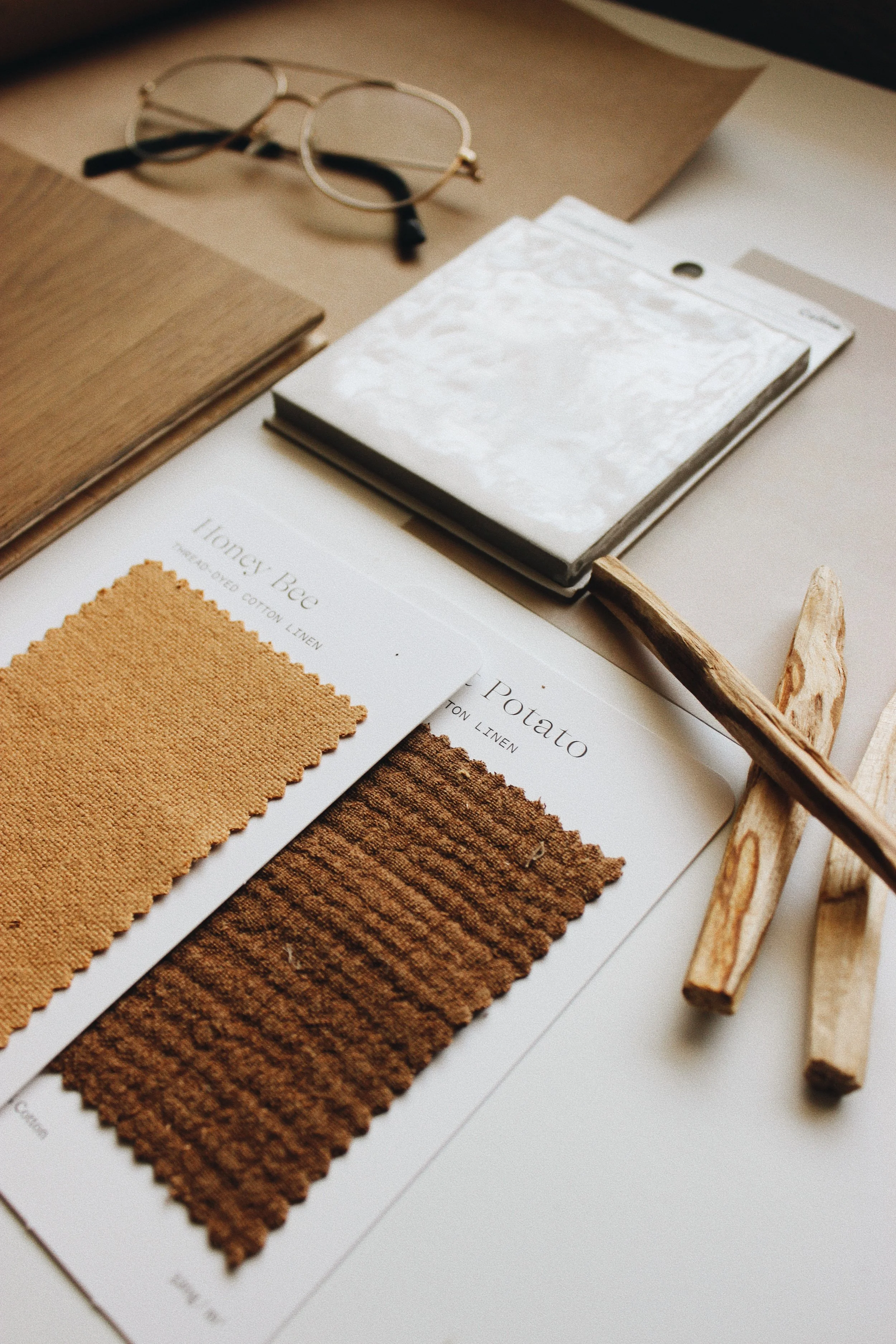 Samples of beige and brown upholstery fabrics, wooden swatches, a pair of eyeglasses, and color swatches labeled "Honey Bee" and "Potato" on a table.