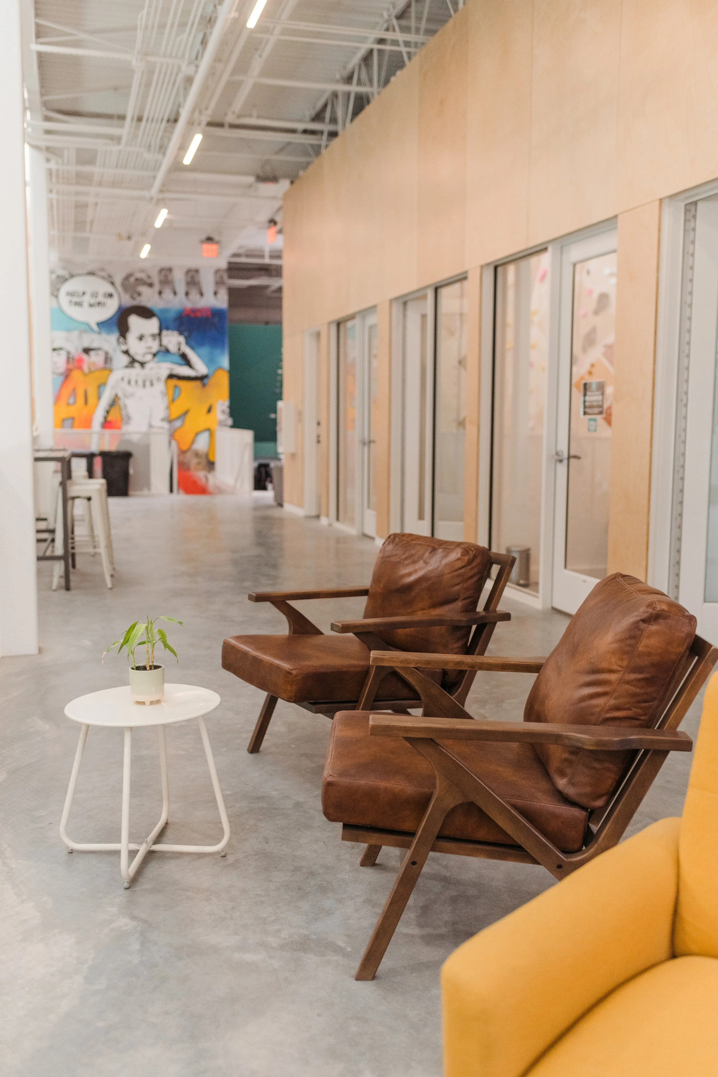 A modern interior with two brown leather armchairs, a small white side table with a plant, yellow seating, and a colorful mural on the wall in the background.